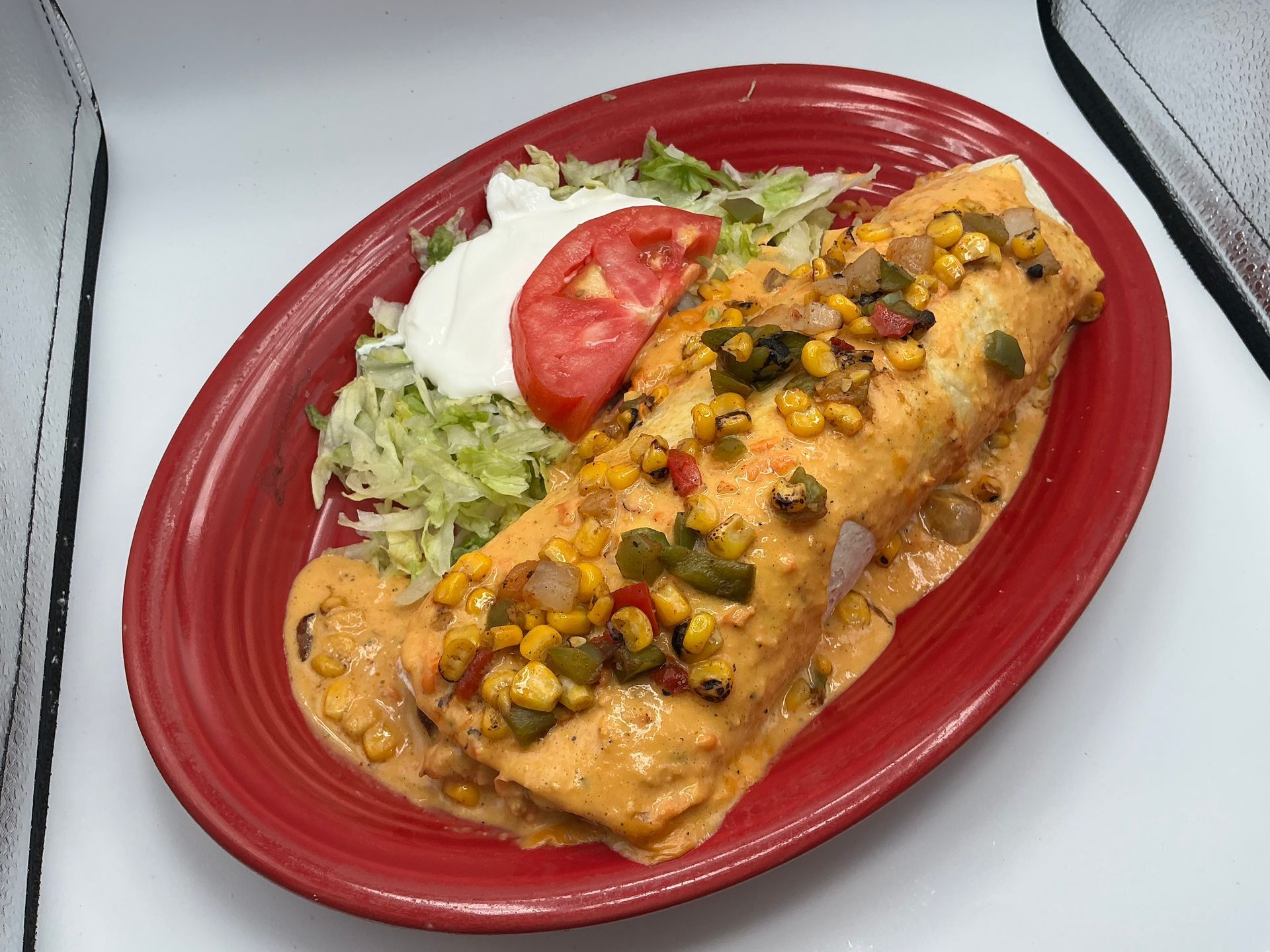 Burrito smothered in sauce with corn, peppers, lettuce, tomato, and sour cream on a red plate.