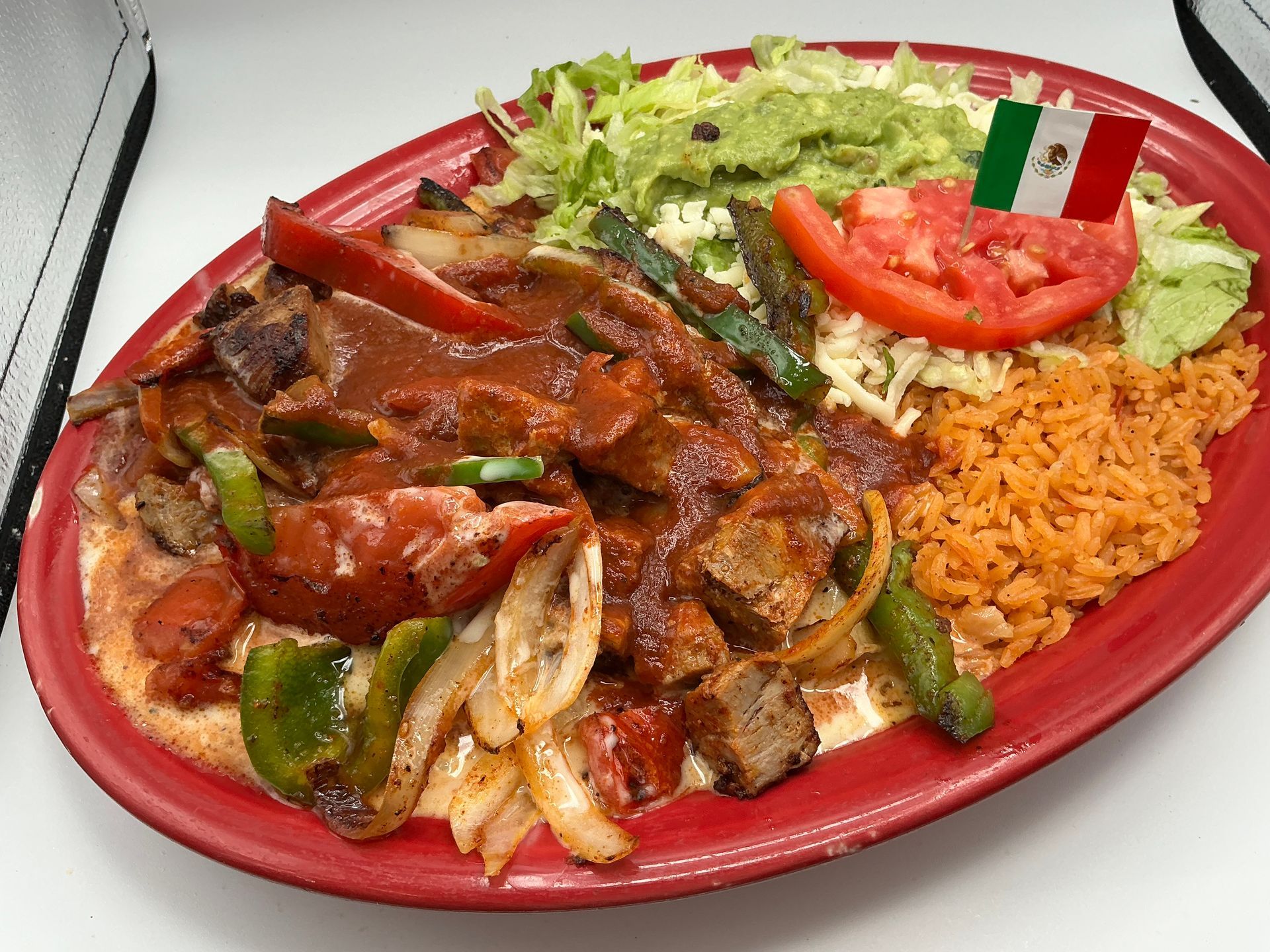 Mexican dish with meat, peppers, onions, rice, guacamole, tomato, lettuce, and a Mexican flag.