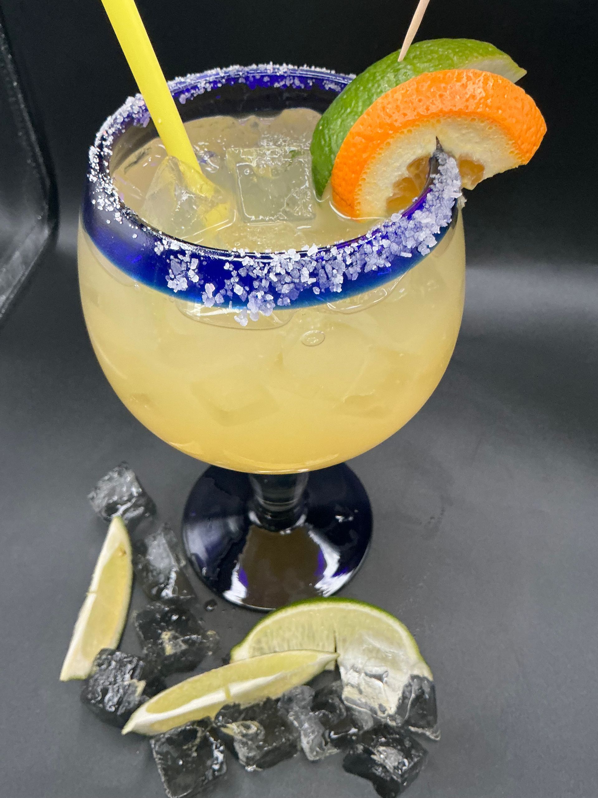 Margarita in a blue-rimmed glass, garnished with citrus slices and a straw, on a black surface with ice and lime wedges.