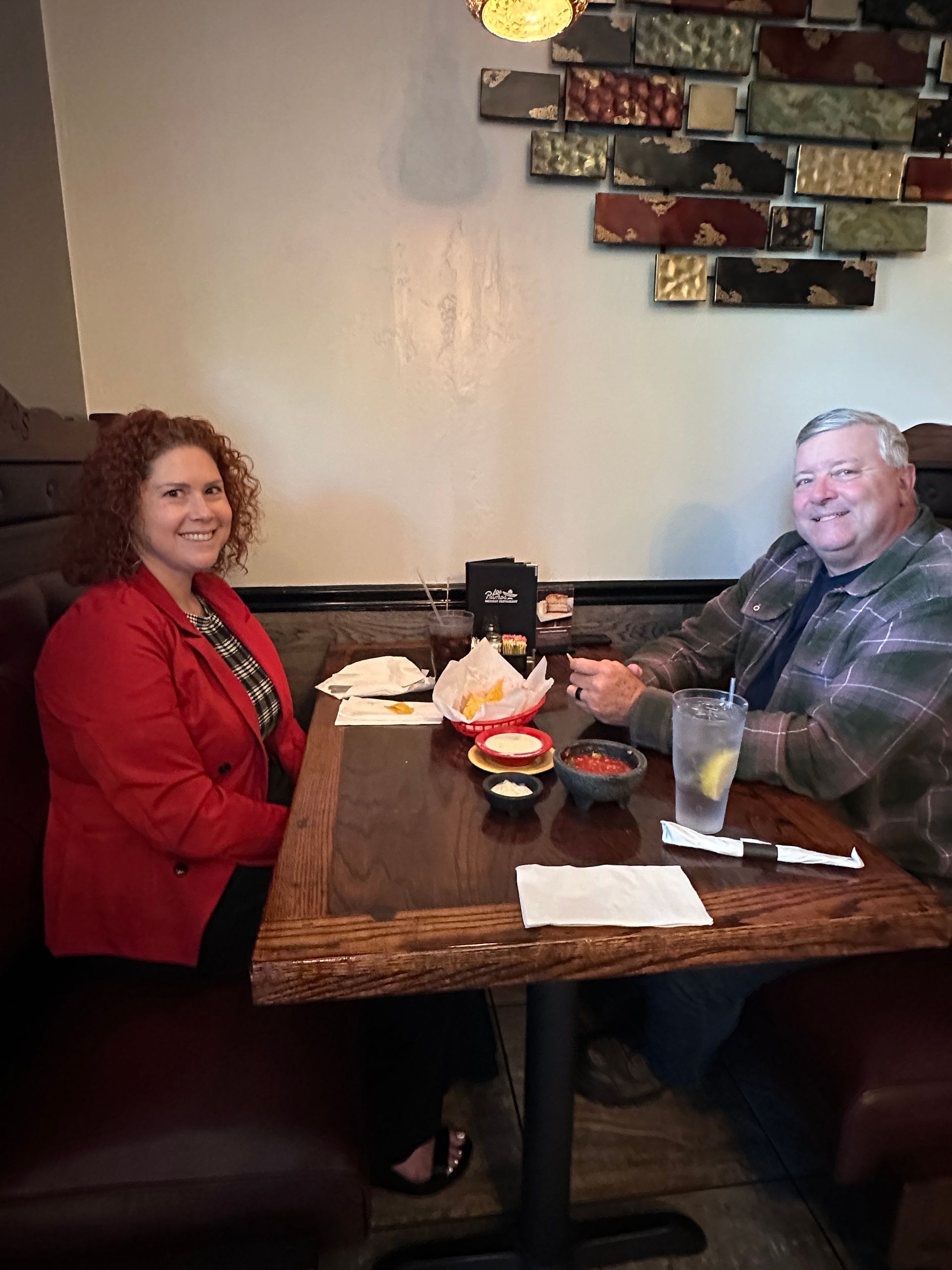 Two people seated at a restaurant table with food and drinks. The woman wears a red blazer and smiles. The man smiles.