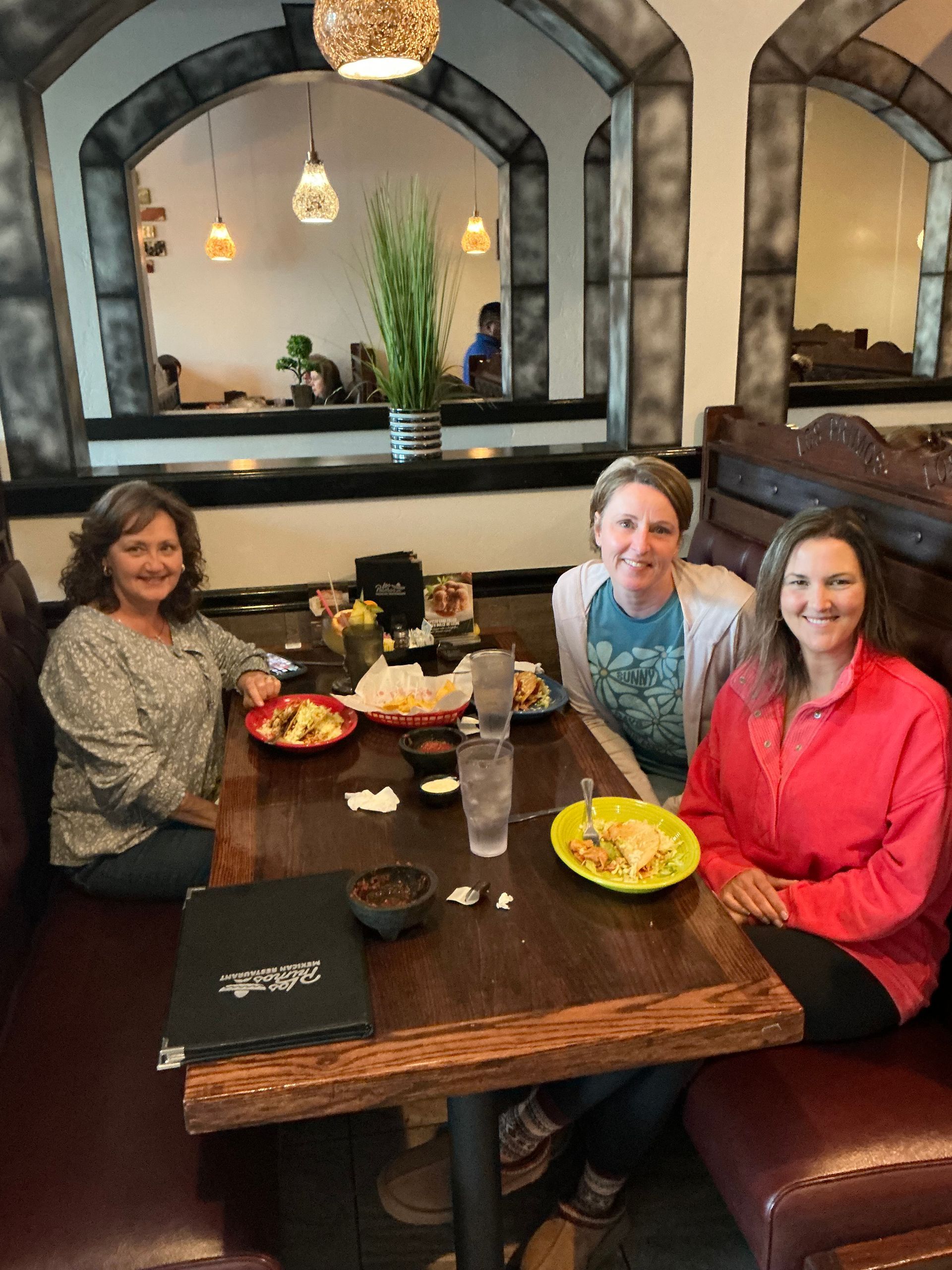 Three people smiling at a restaurant table with food and drinks. Arched mirrors in the background.