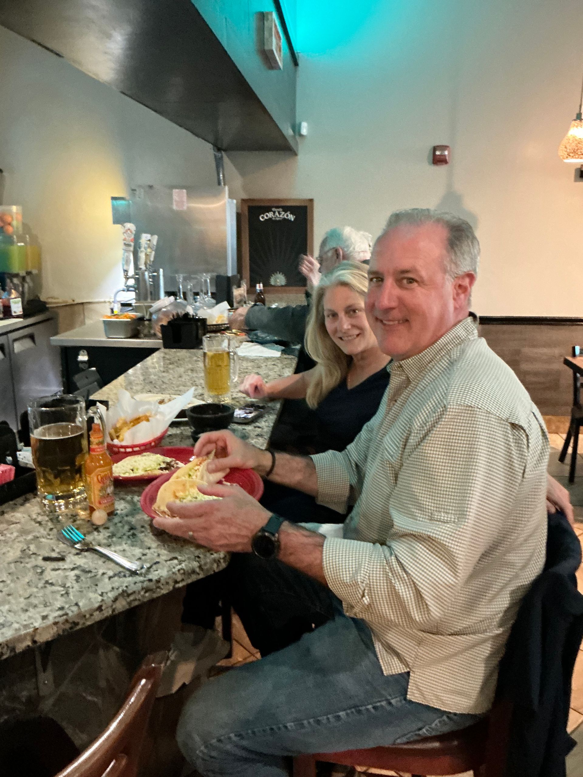 Man and woman at a bar, eating food. Man smiles at camera, holding a taco. The bar has granite counter.