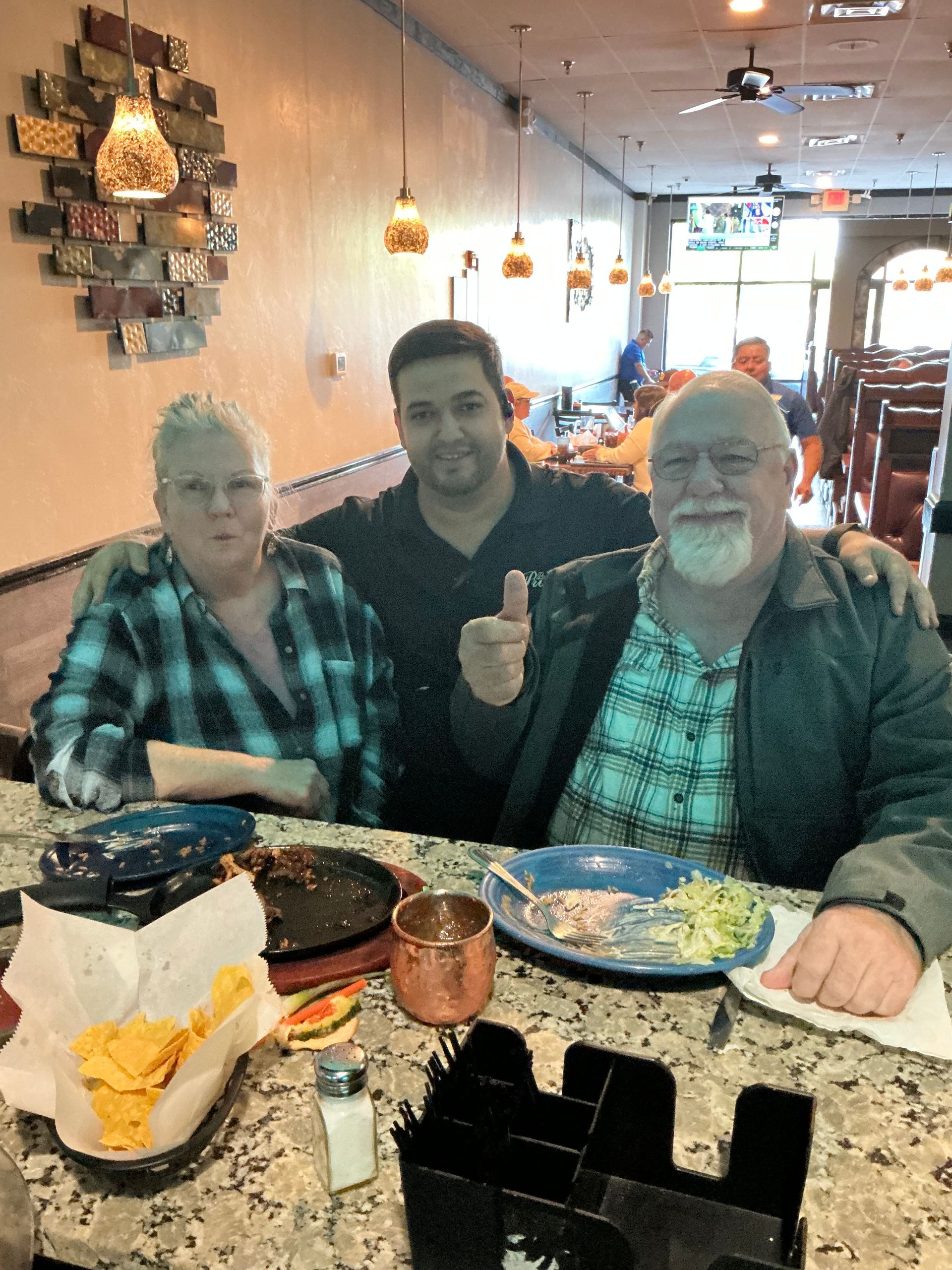 Three people at a restaurant, one giving thumbs up. Food on table; bar setting.