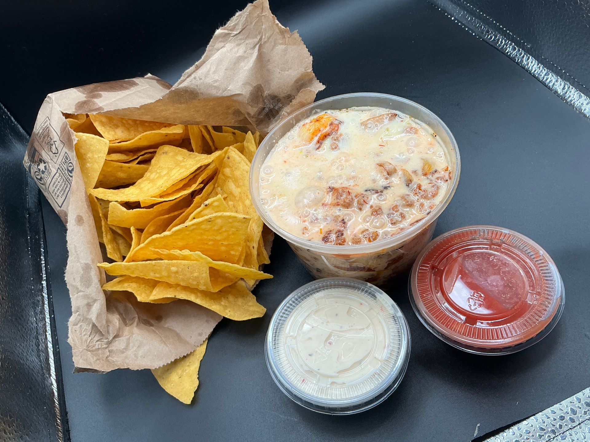 Chips with queso, salsa, and dip in containers. Brown paper bag with chips on a black surface.