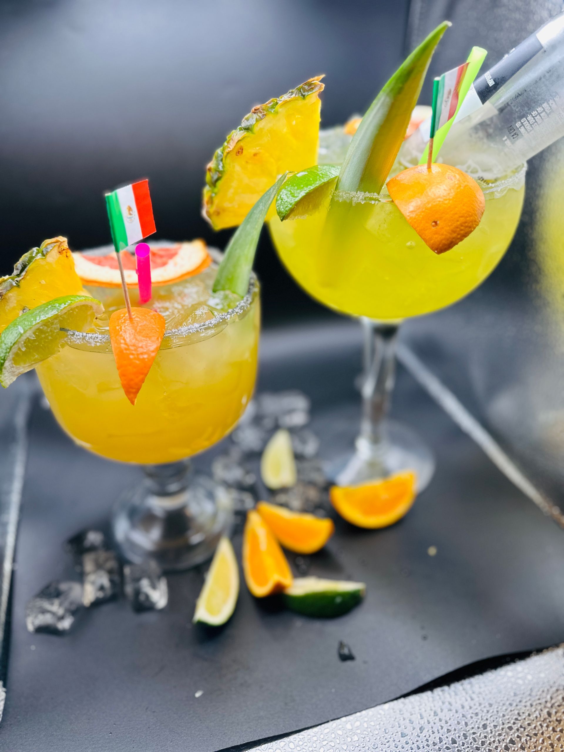 Two margarita cocktails garnished with fruit and flags, on a black surface.