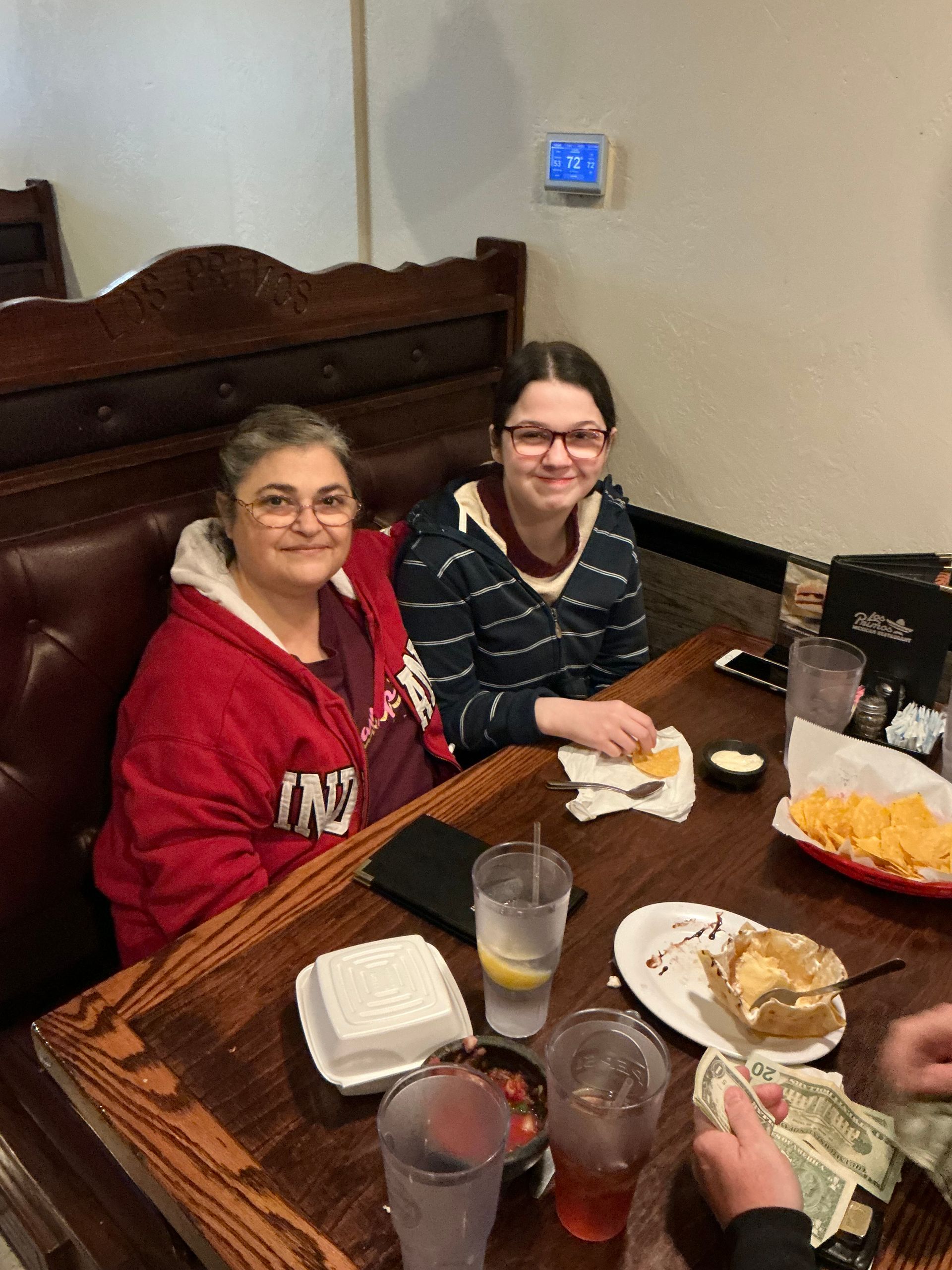 Two people seated at a restaurant table. One in a red hoodie, the other in a striped shirt, both smiling. Food on table.