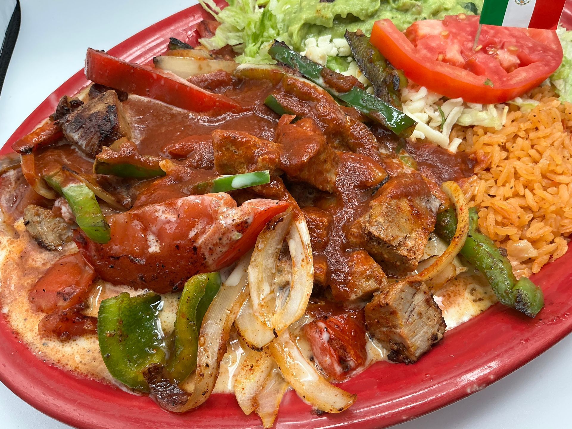 Grilled meat and vegetables in a sauce with rice, lettuce, and guacamole on a red plate.