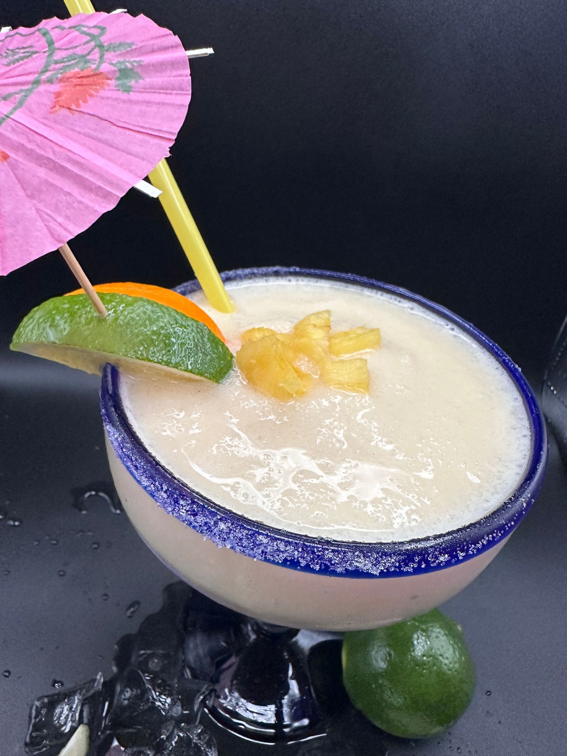 Frozen tropical cocktail in a blue-rimmed glass, garnished with lime, orange, and umbrella.
