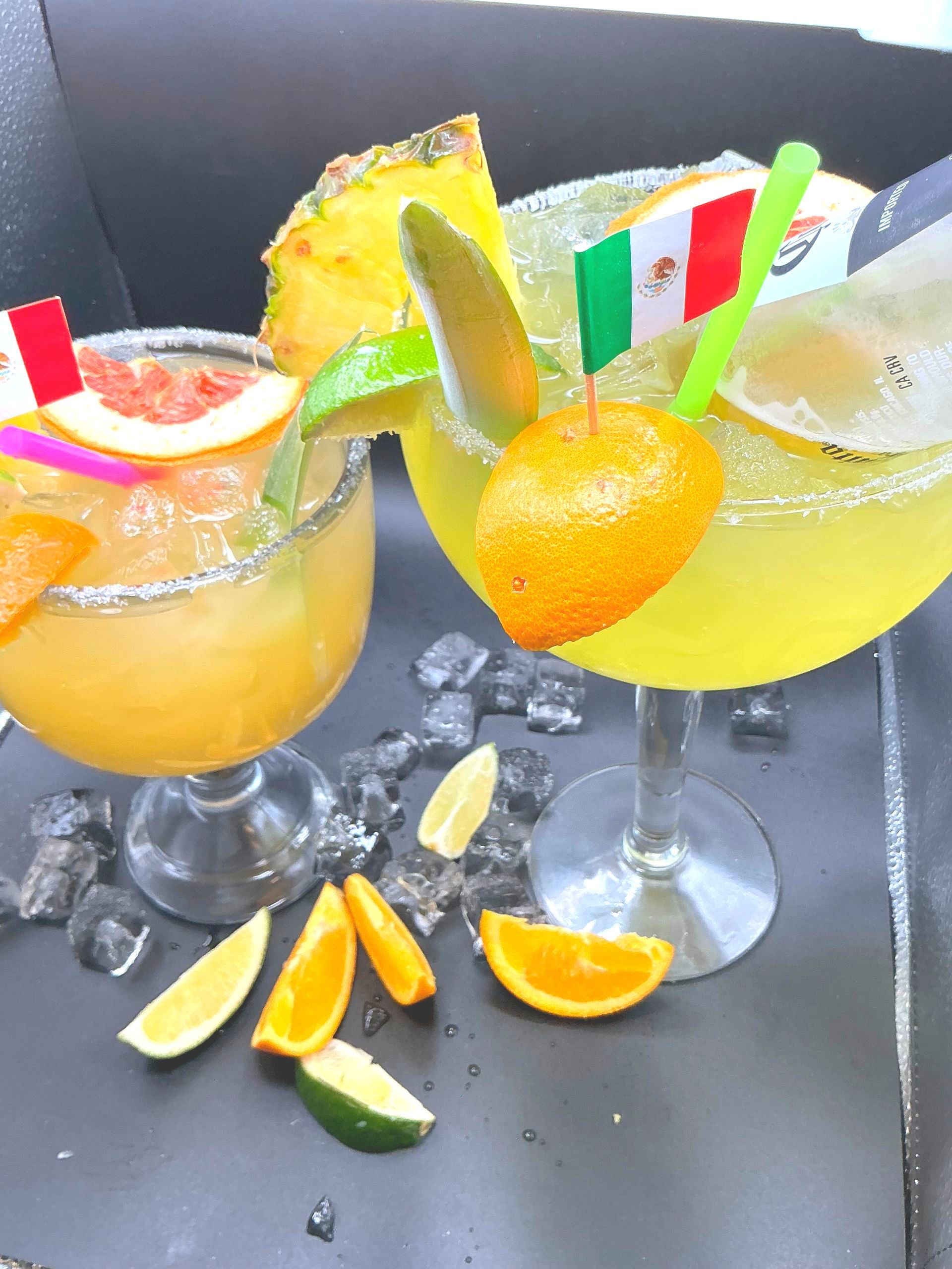 Two colorful margaritas with fruit garnishes, Mexican flags, and ice on a black surface.