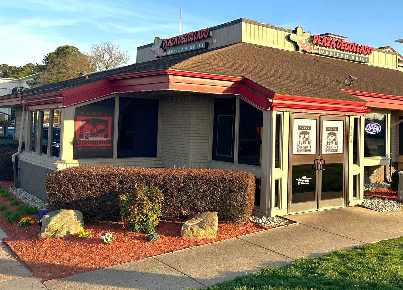Restaurant exterior with red trim and brown roof. Front entrance with two doors. Landscaping includes bushes, rocks, and red mulch.