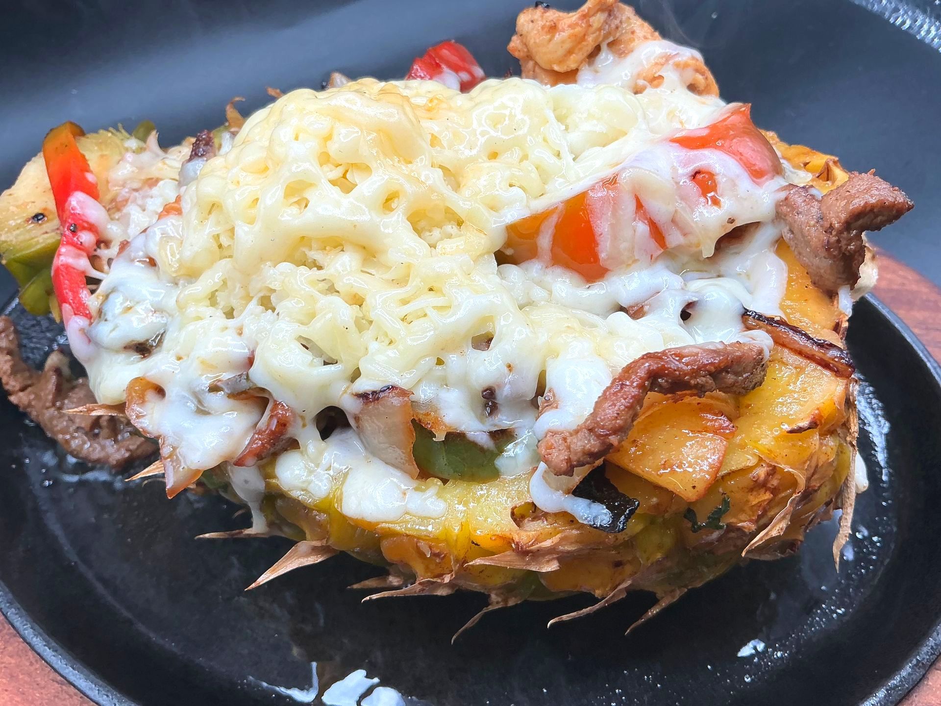 Grilled pineapple bowl filled with meat, vegetables, and melted cheese, served on a black skillet.