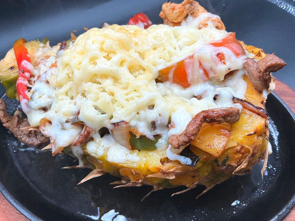 Grilled pineapple bowl filled with meat, vegetables, and melted cheese, served on a black skillet.
