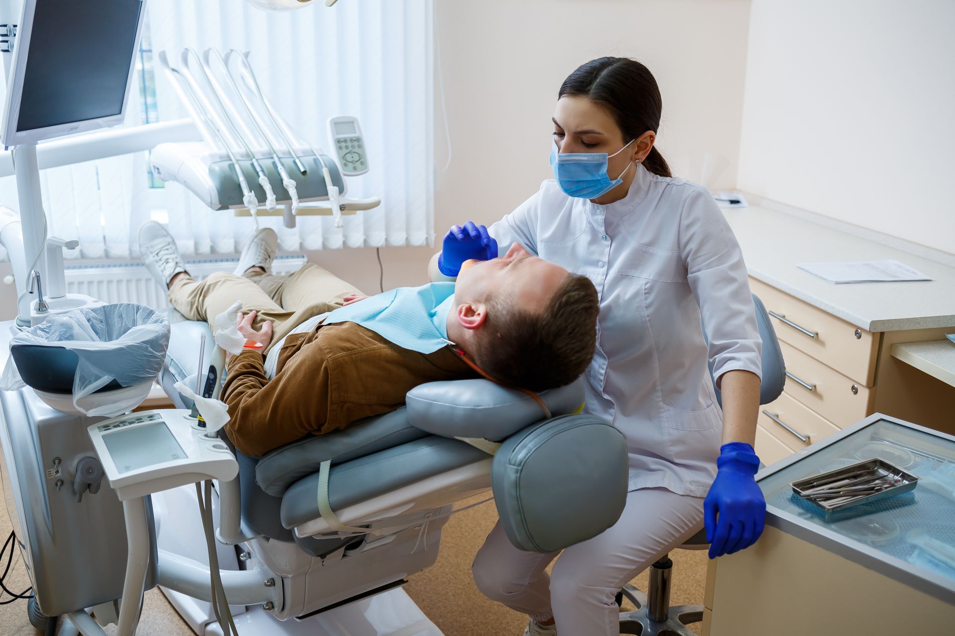 A dentist is examining a patient in a chair with tools, light, and equipment in a clinic room