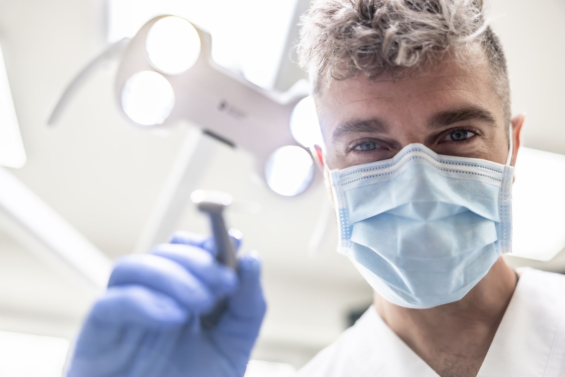 Dentist with mask and gloves holding tool under exam light.