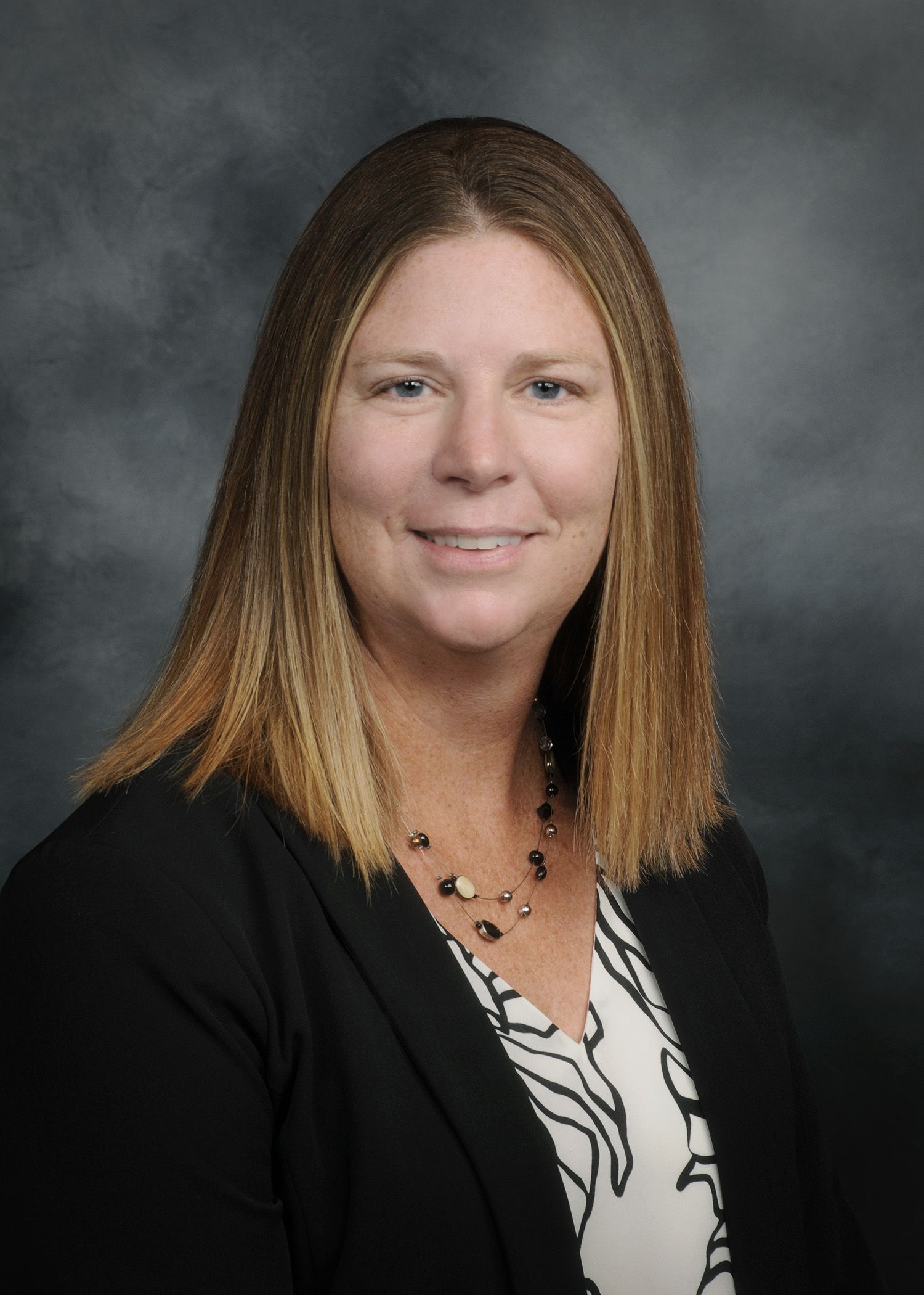 Portrait of a woman in a black blazer and patterned blouse against a gray studio backdrop