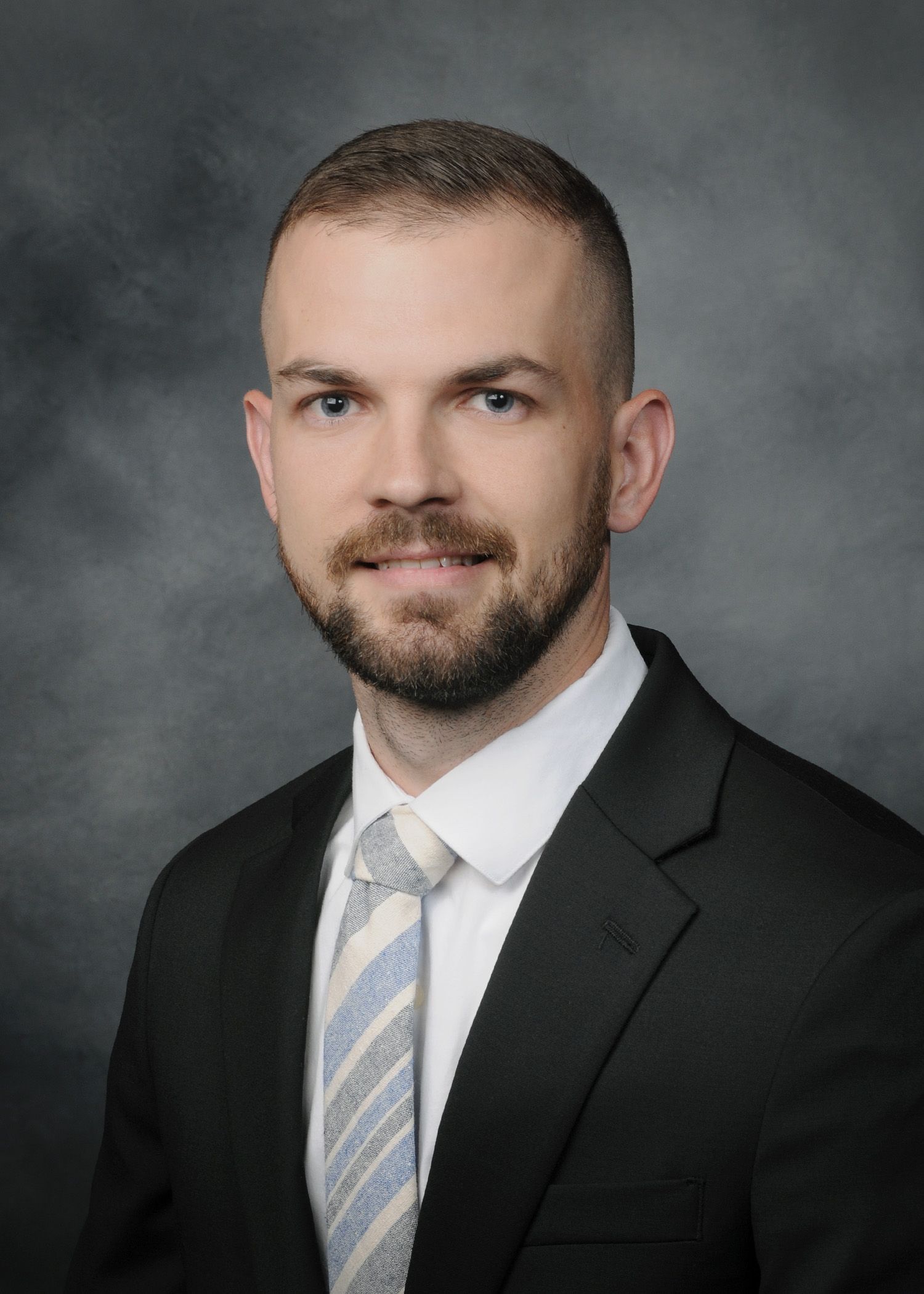 Studio headshot of a man in a dark suit, white shirt, and striped tie against a gray background