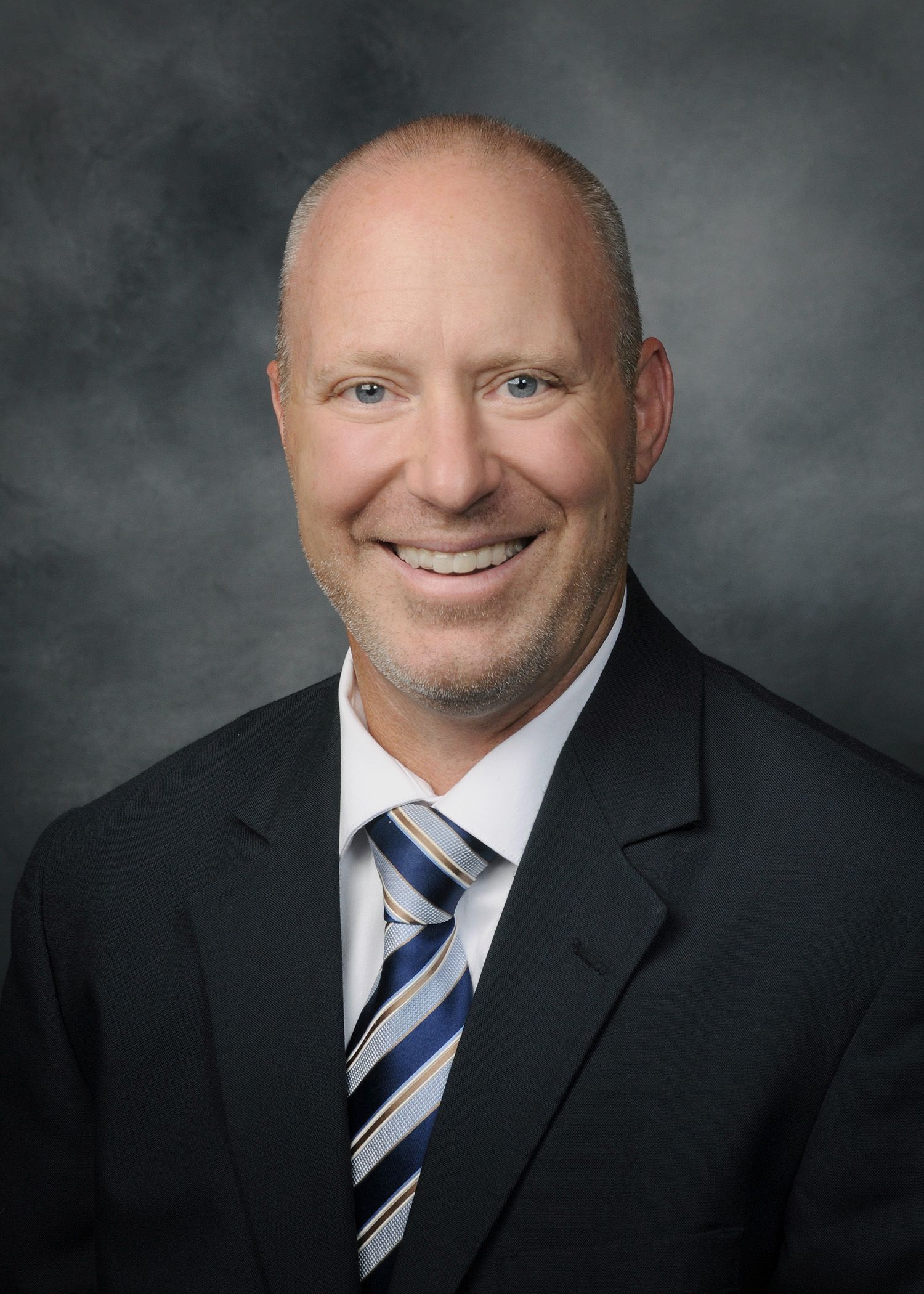 Smiling man in a dark suit and striped tie against a gray studio background