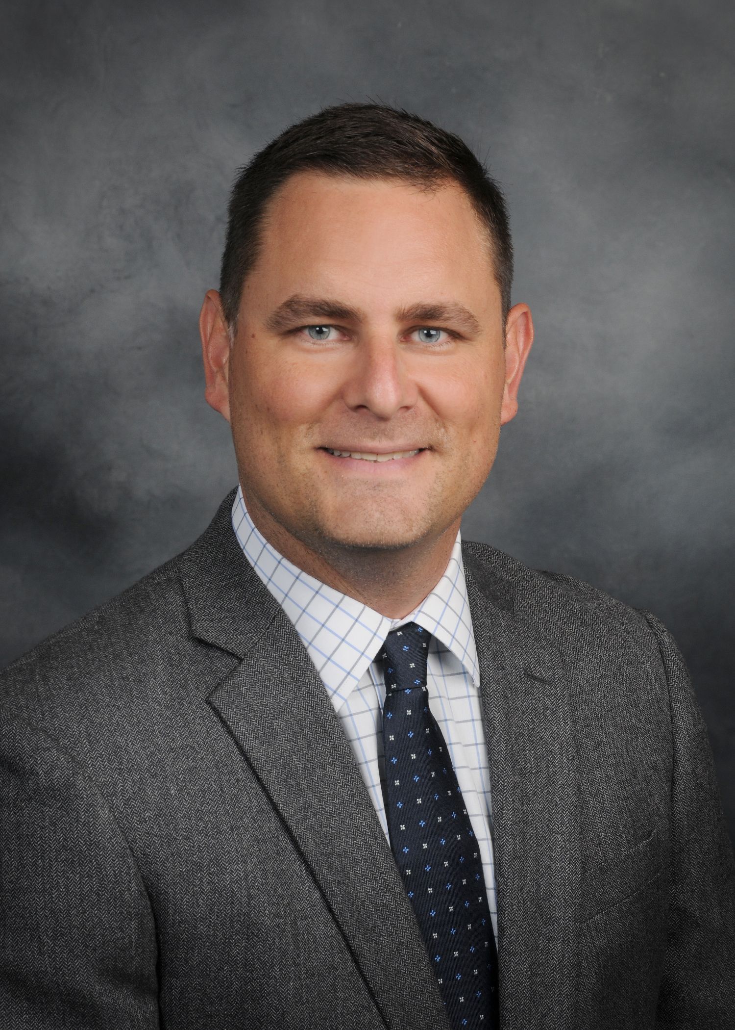 Headshot of a man in a beige suit and blue tie against a gray studio background