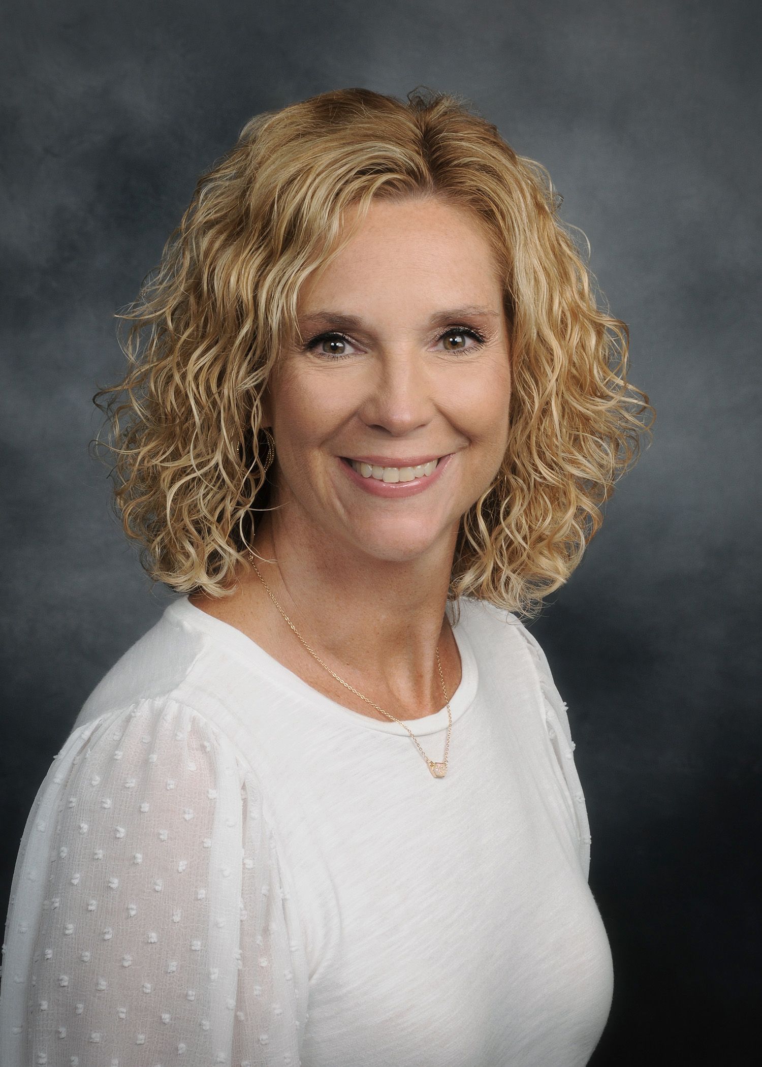 Portrait of a smiling woman with curly blonde hair in a white top against a gray backdrop