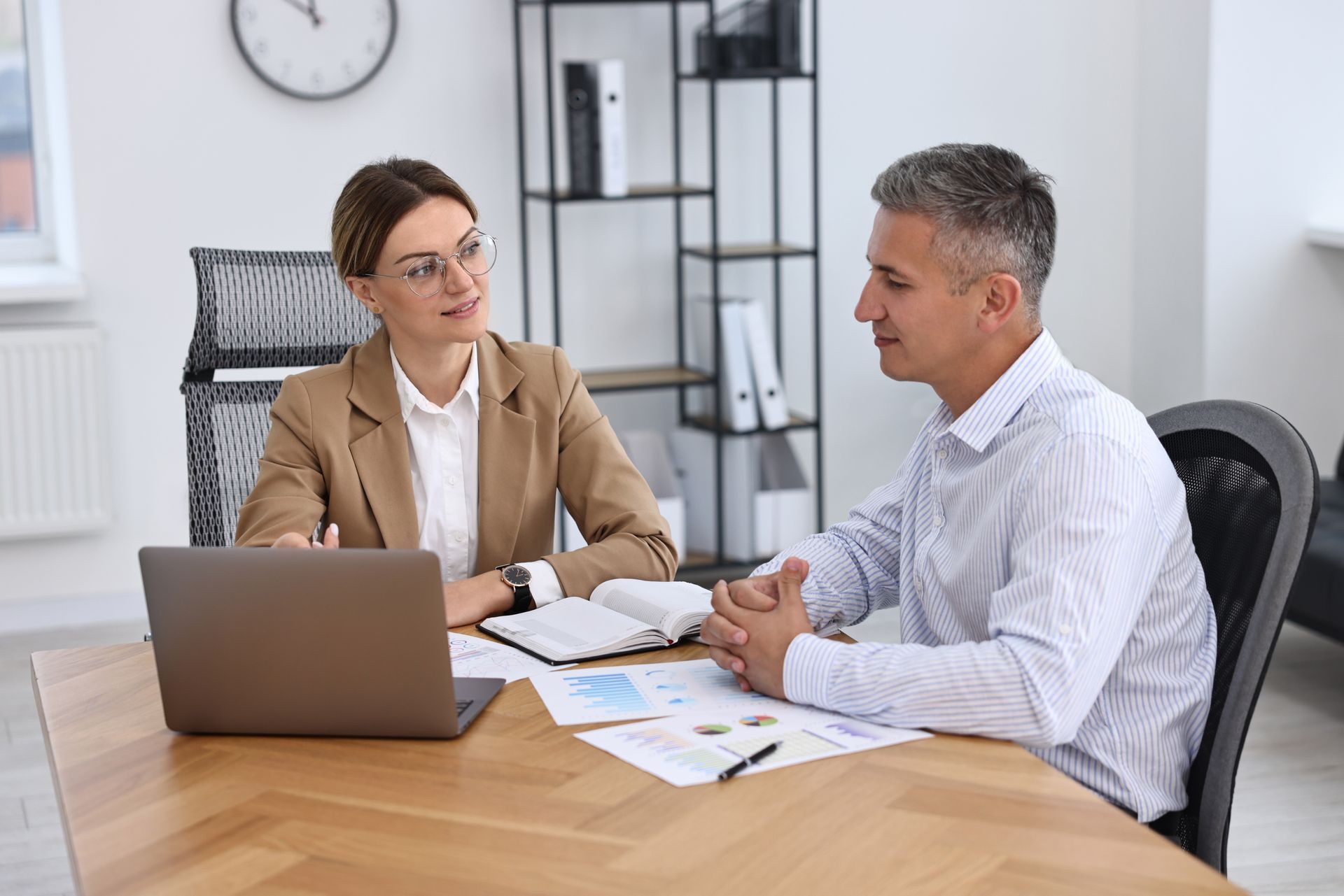 Two coworkers discussing documents at a desk with a laptop in a bright office