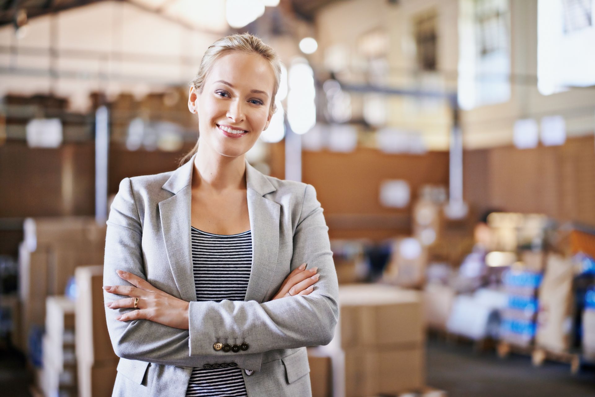 Smiling woman in a gray blazer with arms crossed in a warehouse setting