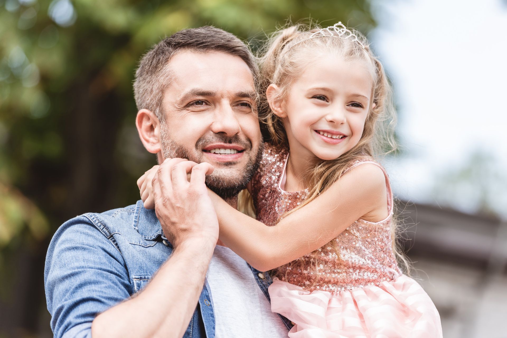Smiling man carrying a smiling child outdoors, both in casual clothes.