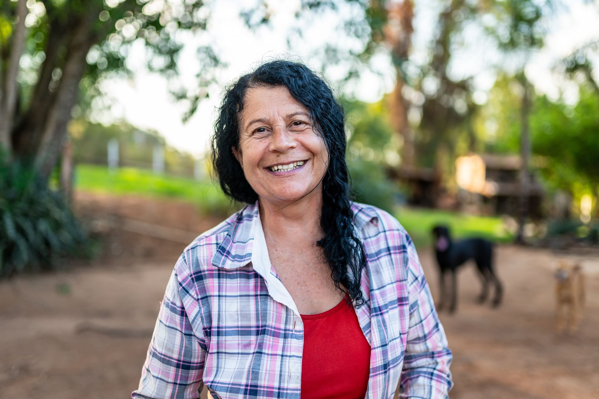Smiling woman in a plaid shirt and red top standing outdoors with trees and a dog in the background