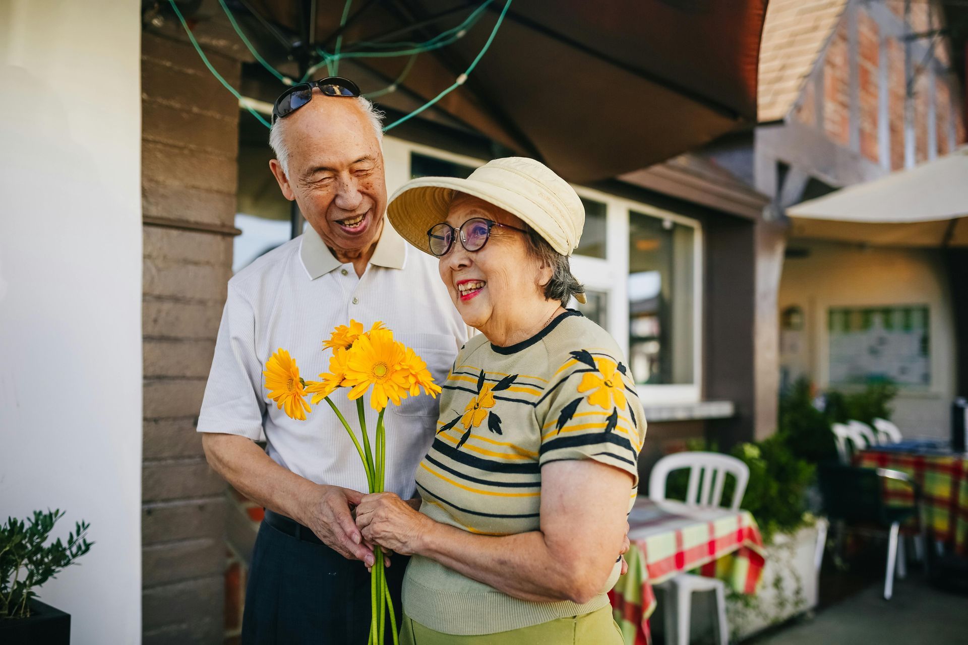 Two people smiling and holding yellow flowers outside a house