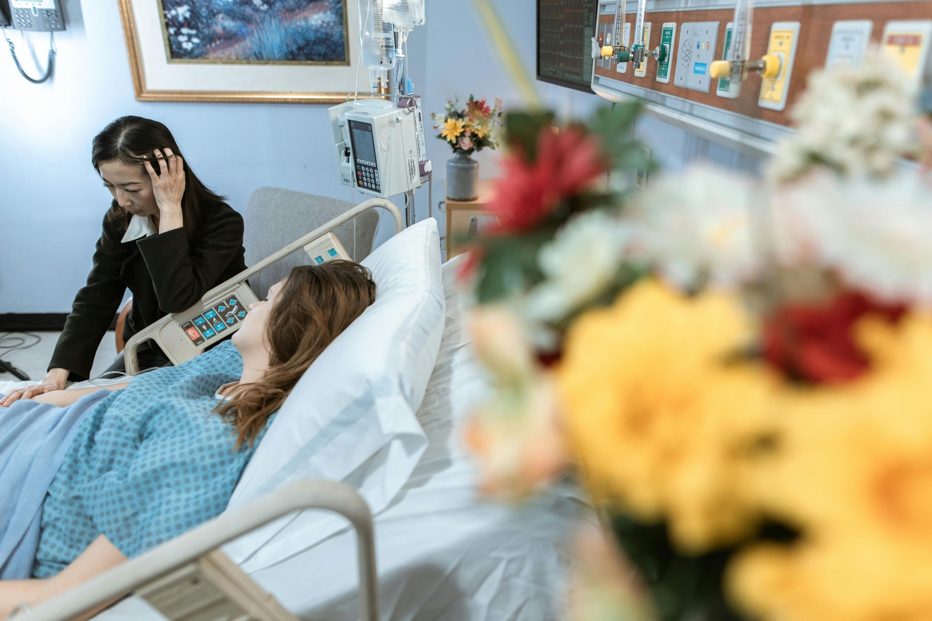 Hospital room with a woman sitting beside a patient resting in bed, flowers blurred in the foreground.
