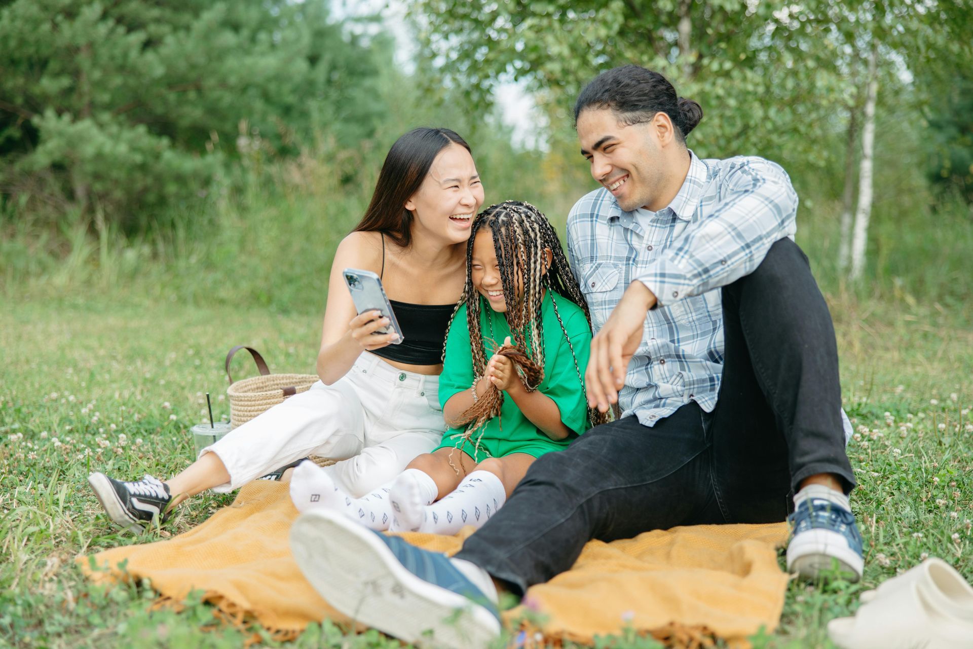 Family sitting on a blanket in a grassy park, smiling and looking at a phone.