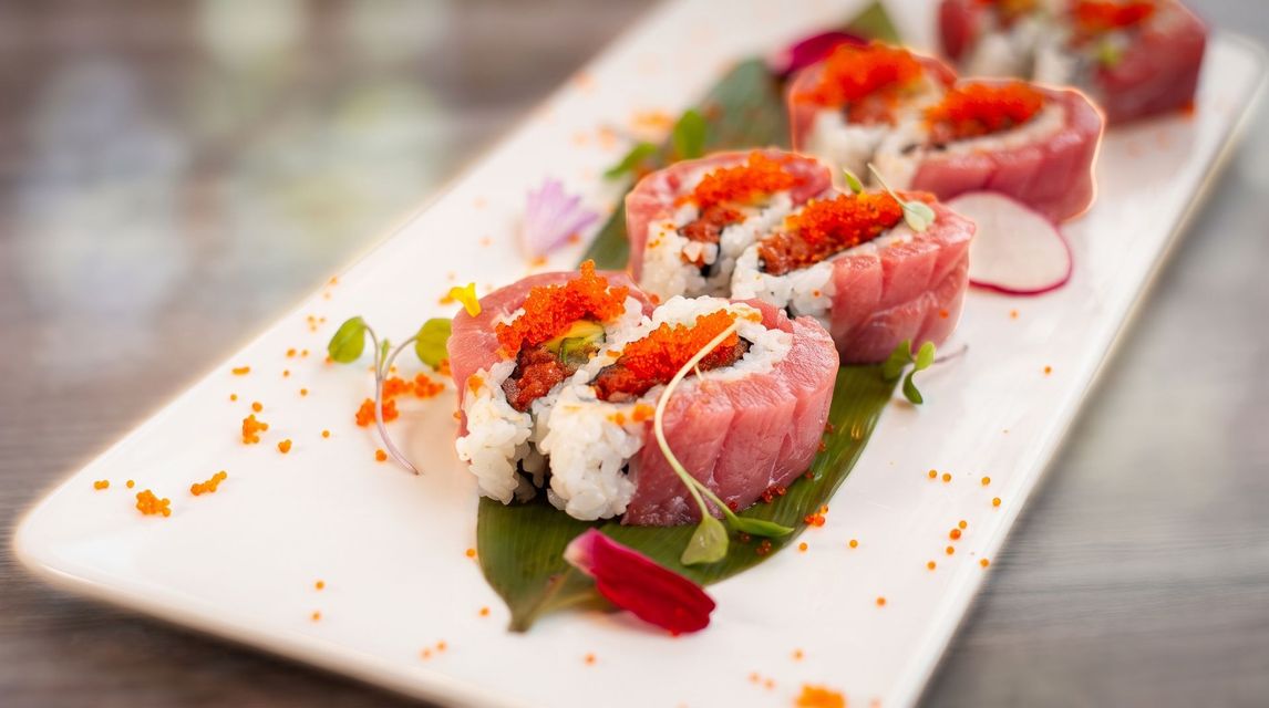 Sushi rolls topped with pink fish and orange roe on a white rectangular plate, garnished with greens