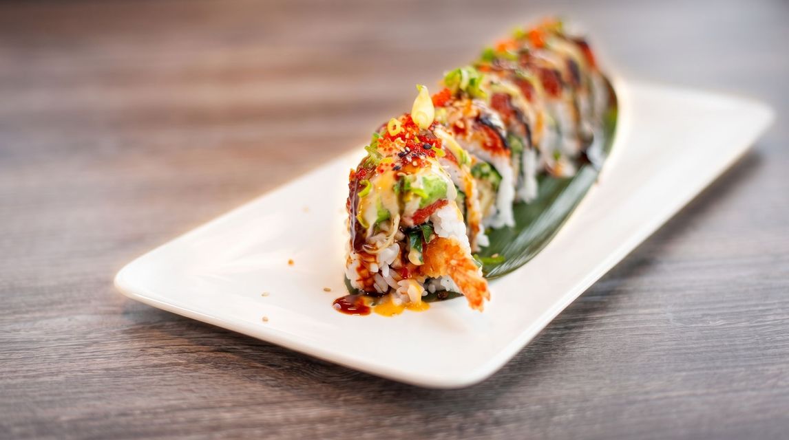 Sushi roll topped with avocado, sauce, and spices on a white rectangular plate