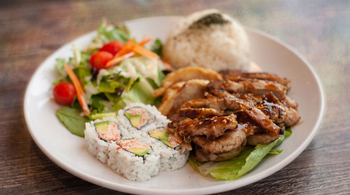 Plate with grilled chicken, sushi roll, salad, and rice on a table