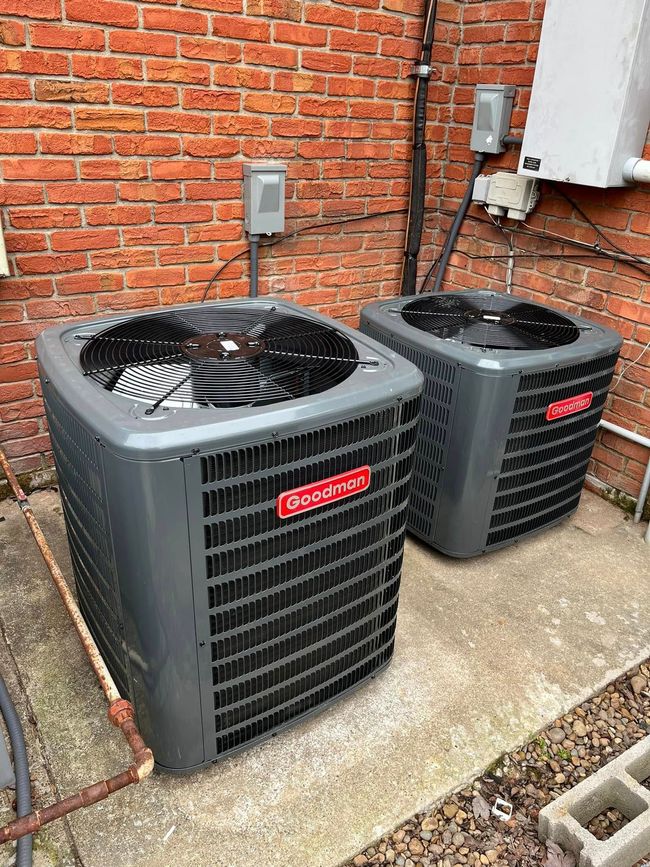 Two gray outdoor HVAC units beside a brick wall on a concrete pad, one larger with a visible fan.