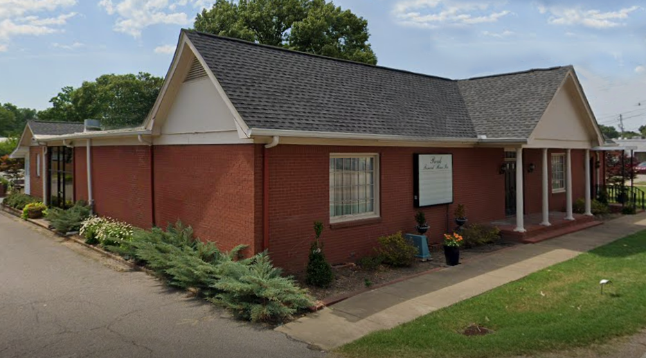 Single-story red brick building with white trim, gray roof, and a small porch along a paved drive.