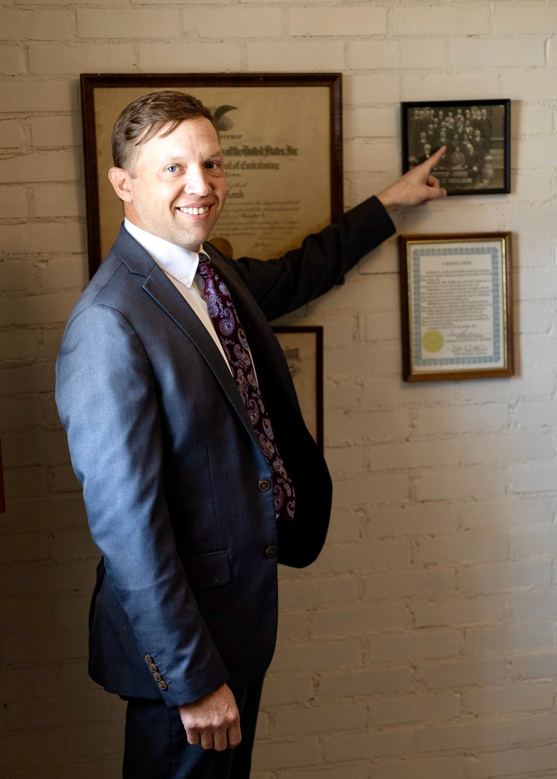 Man in a suit pointing to a framed plaque on a brick wall next to certificates