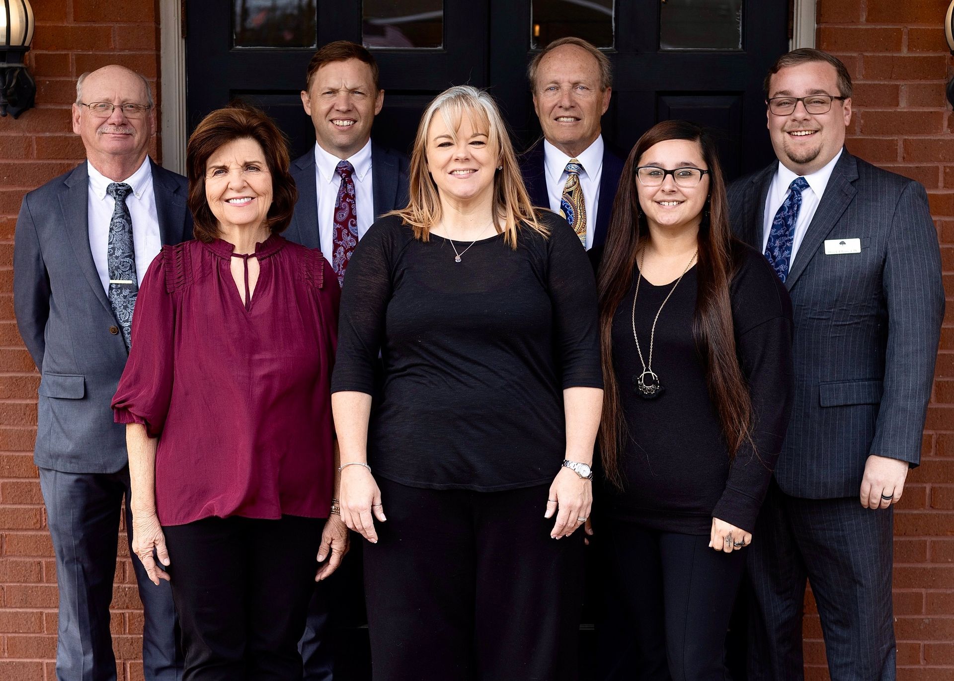 Group of seven professionals posing in business attire outdoors in front of a brick wall