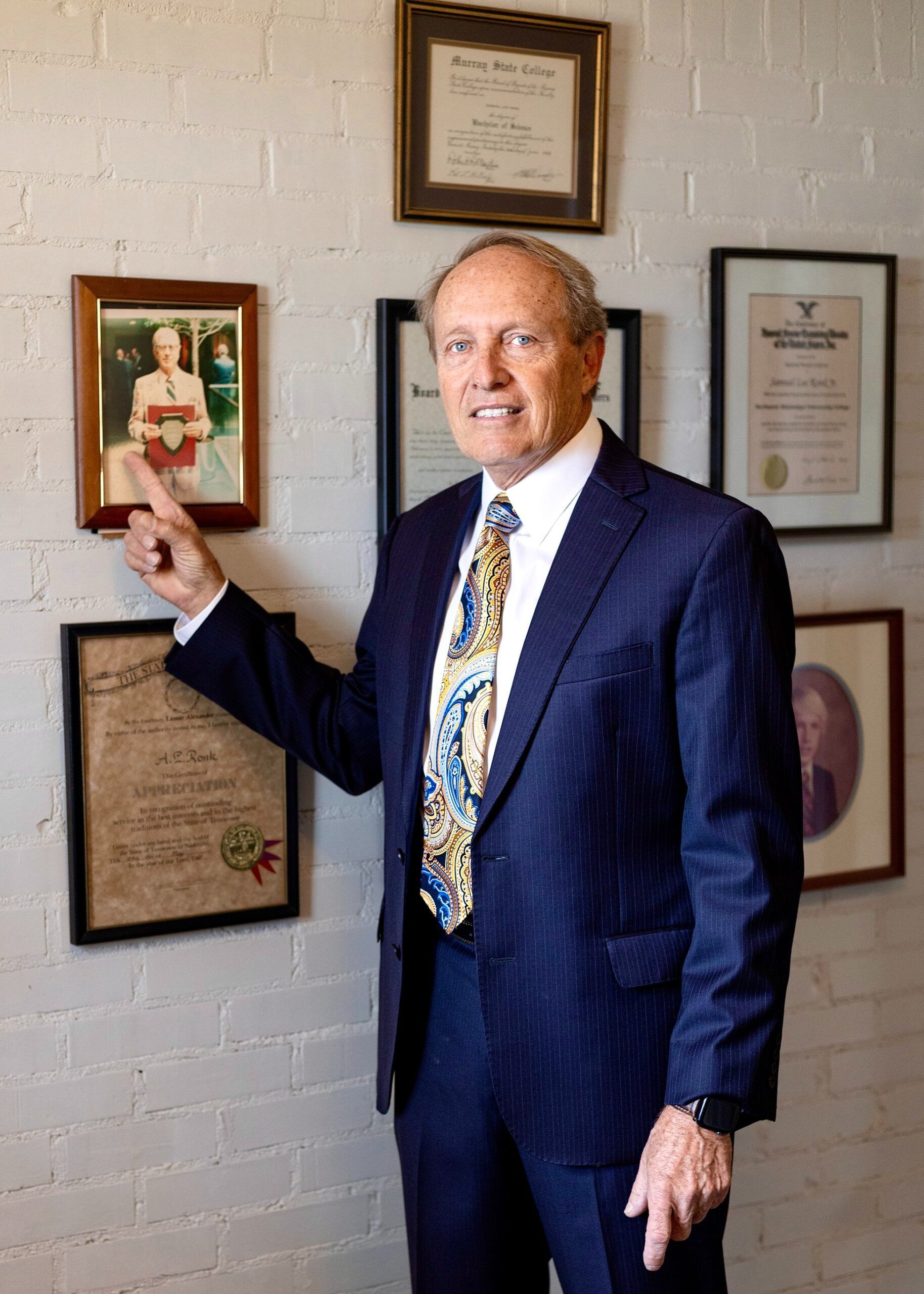 Man in a blue suit standing by framed certificates on a white brick wall, pointing at one frame