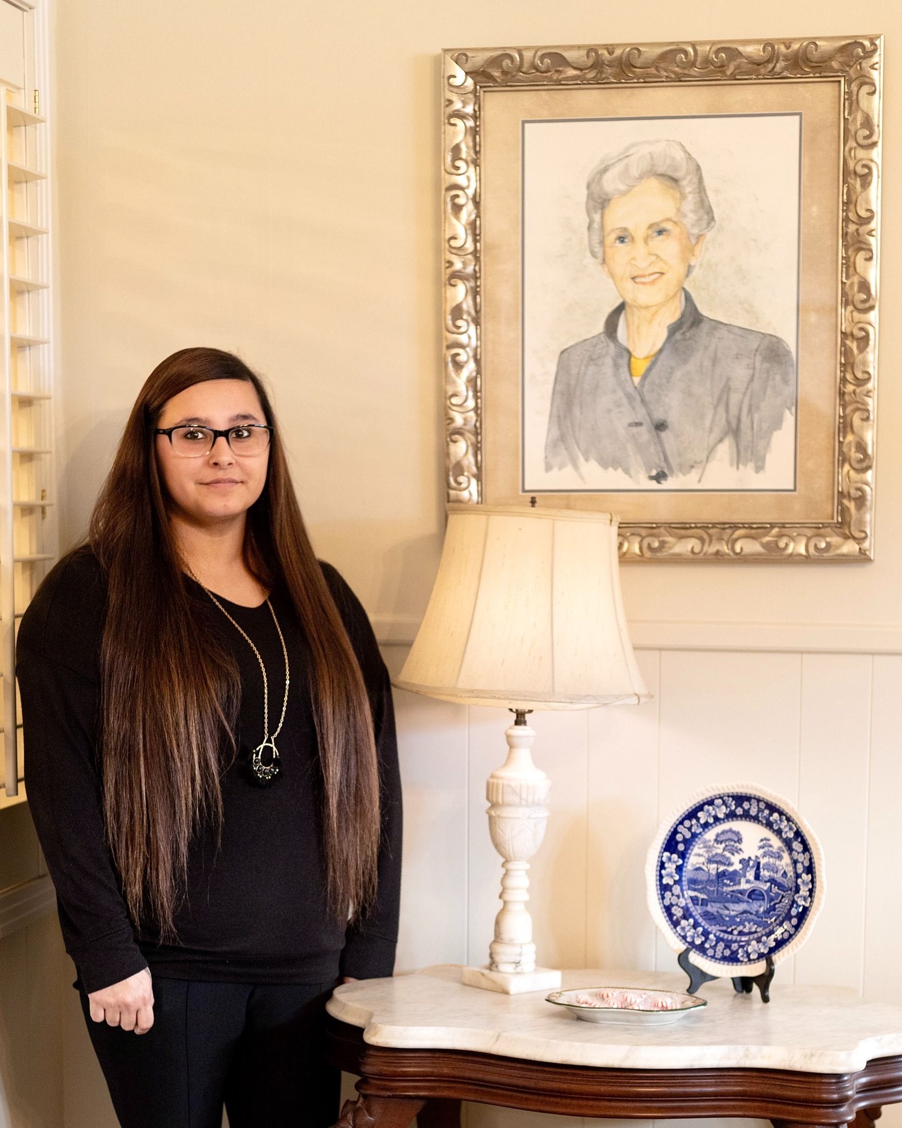Woman standing beside a table lamp under a framed portrait in a softly lit room