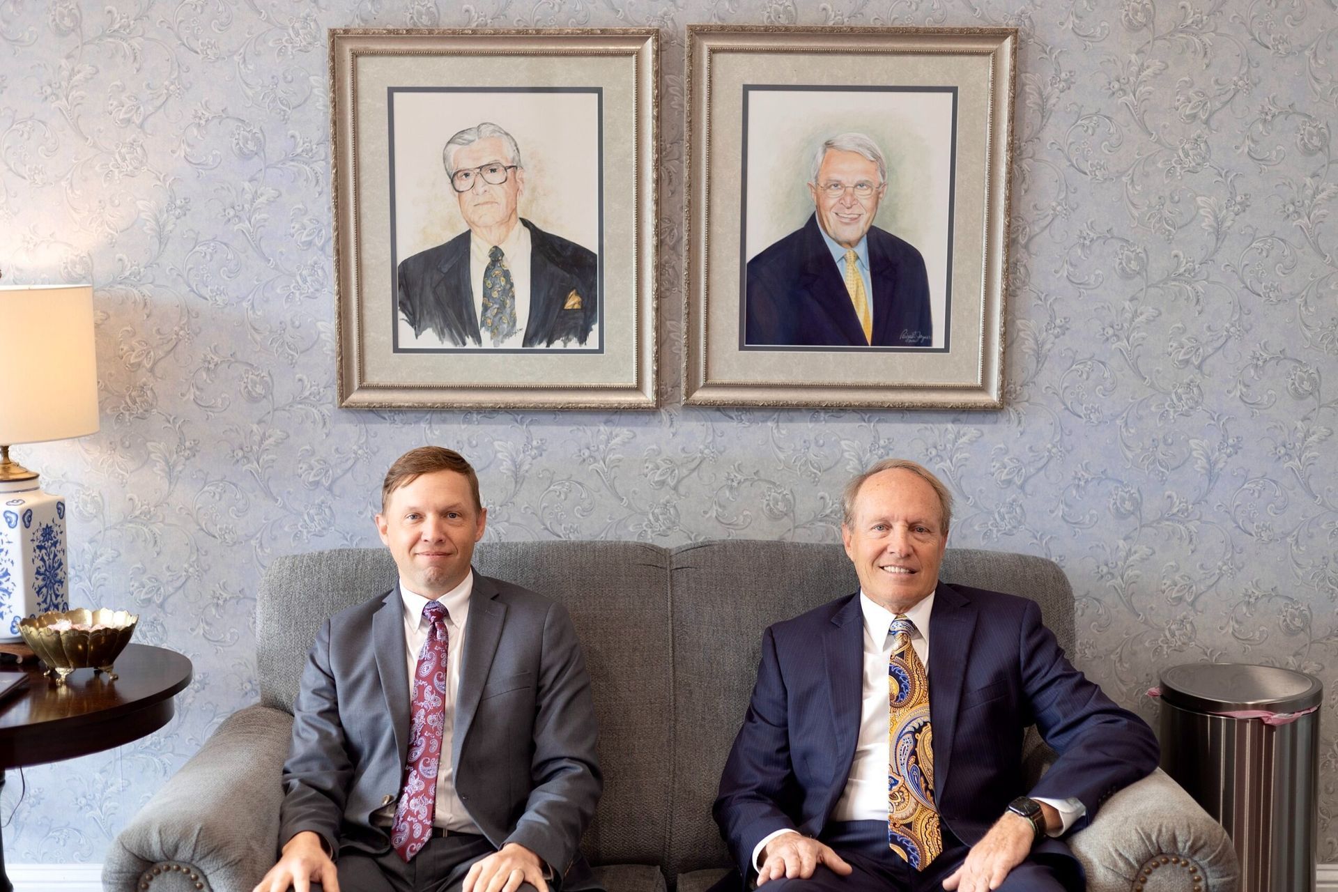 Two men seated on a gray sofa beneath framed portraits in a formal room.