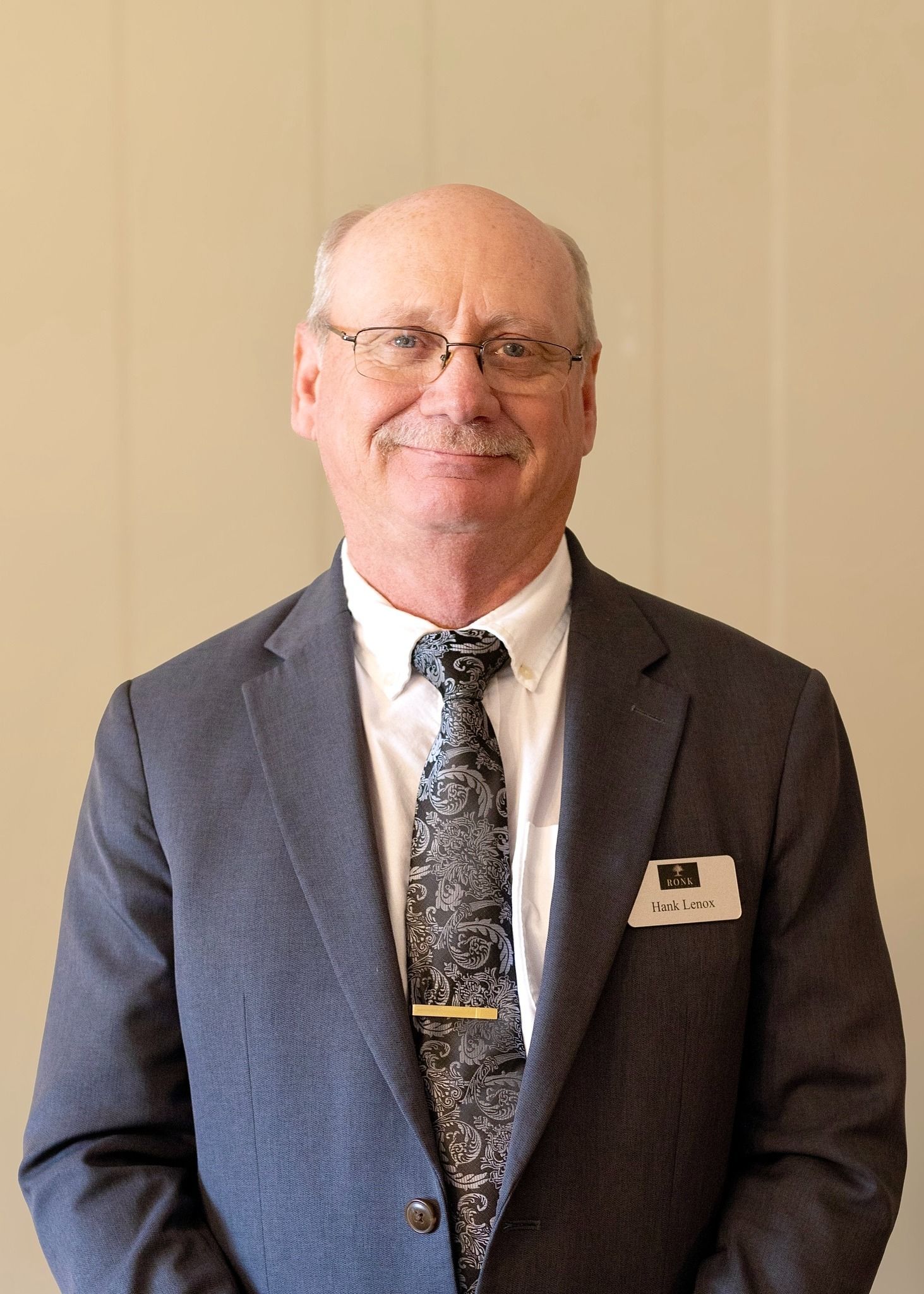 Smiling man in a gray suit and patterned tie, wearing a name badge, standing against a beige wall.