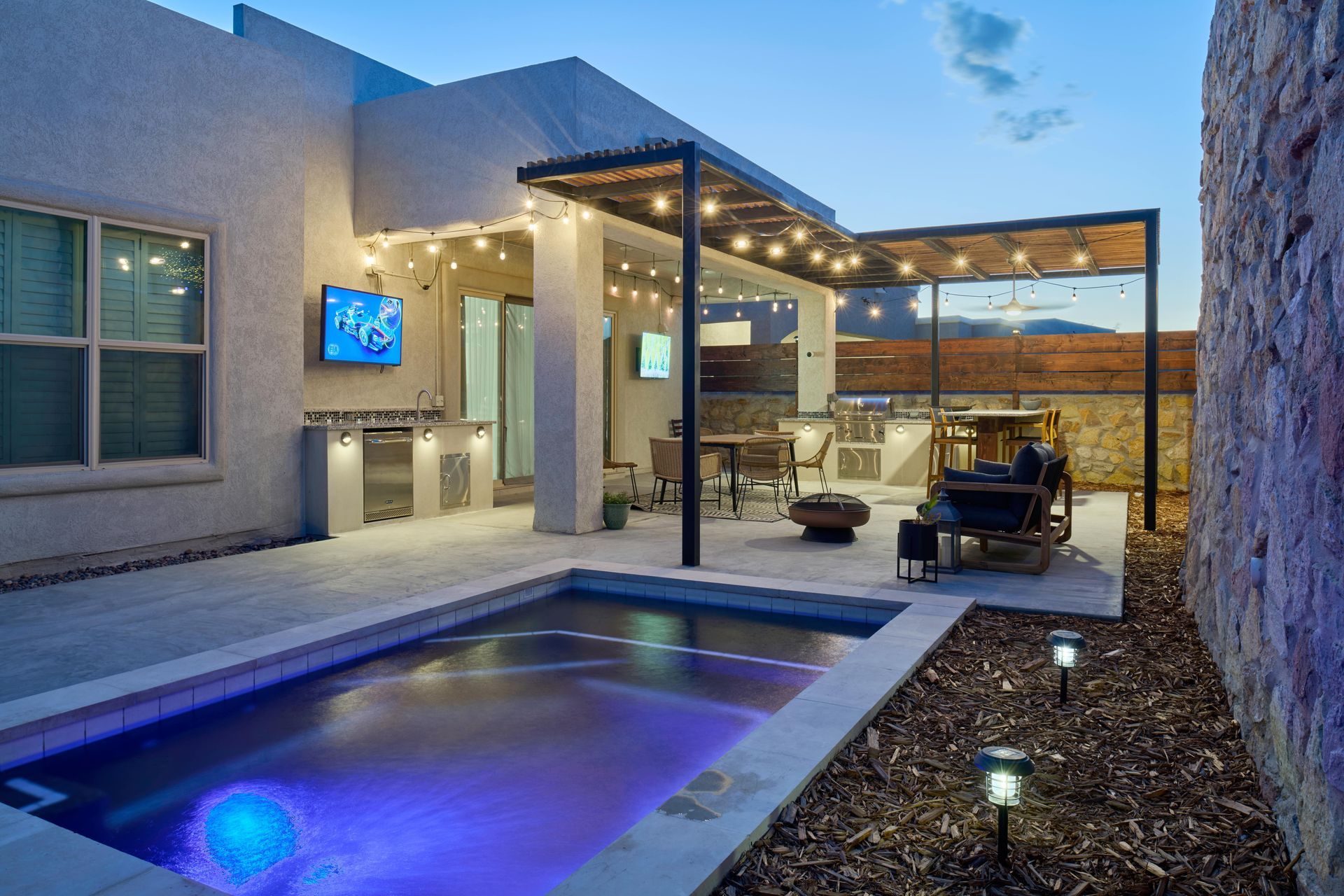 Outdoor patio with pool, kitchen, and seating area illuminated by string lights.