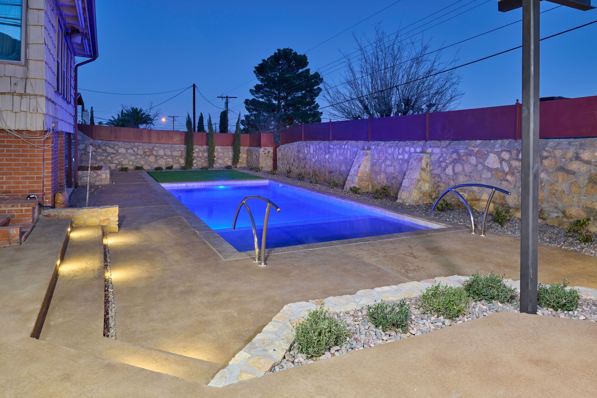 A glowing rectangular swimming pool in a stone-walled backyard at dusk, featuring stepped landscaping and lighting.