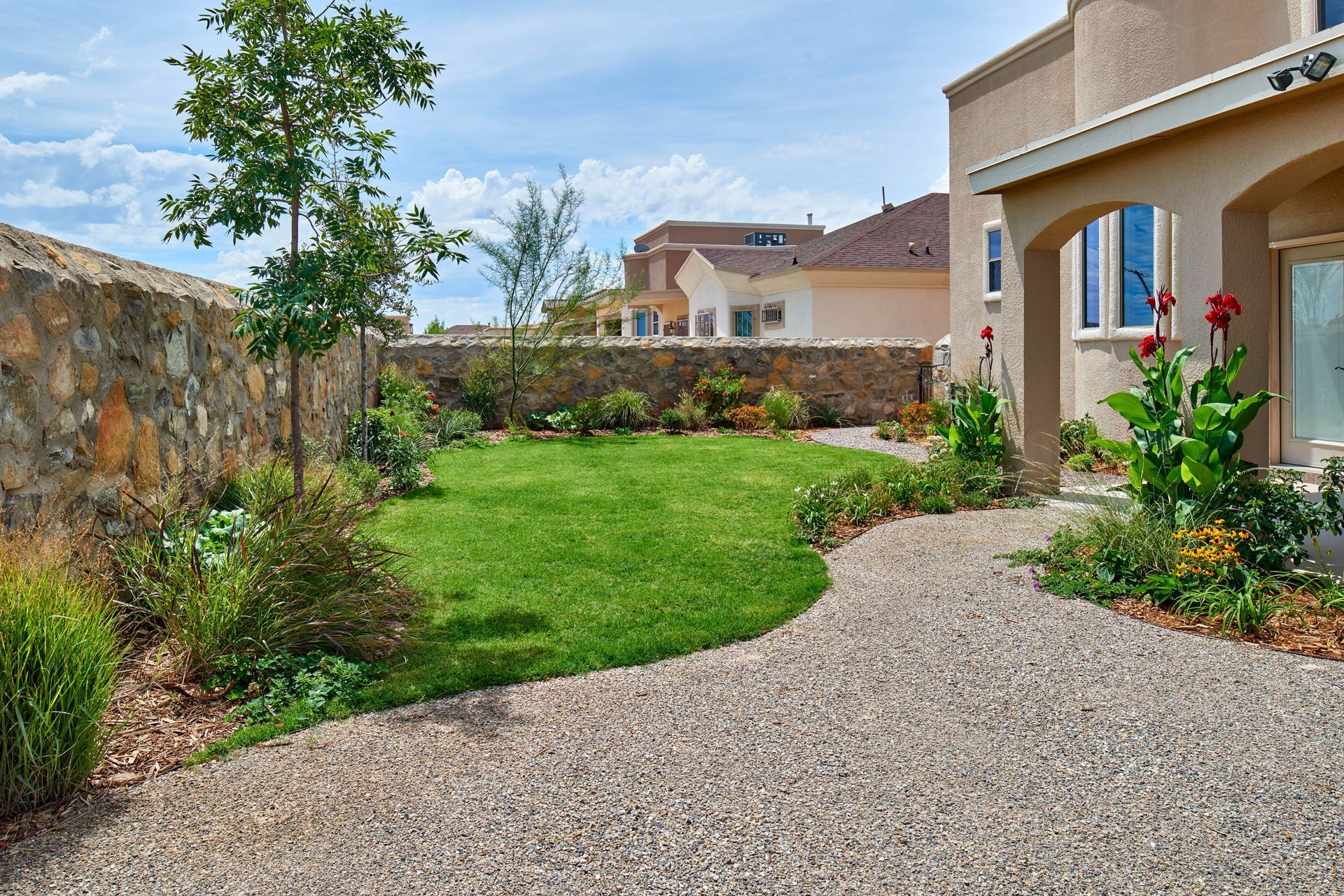 Gravel path leads past lawn and garden with a stone wall and beige house under a cloudy sky.