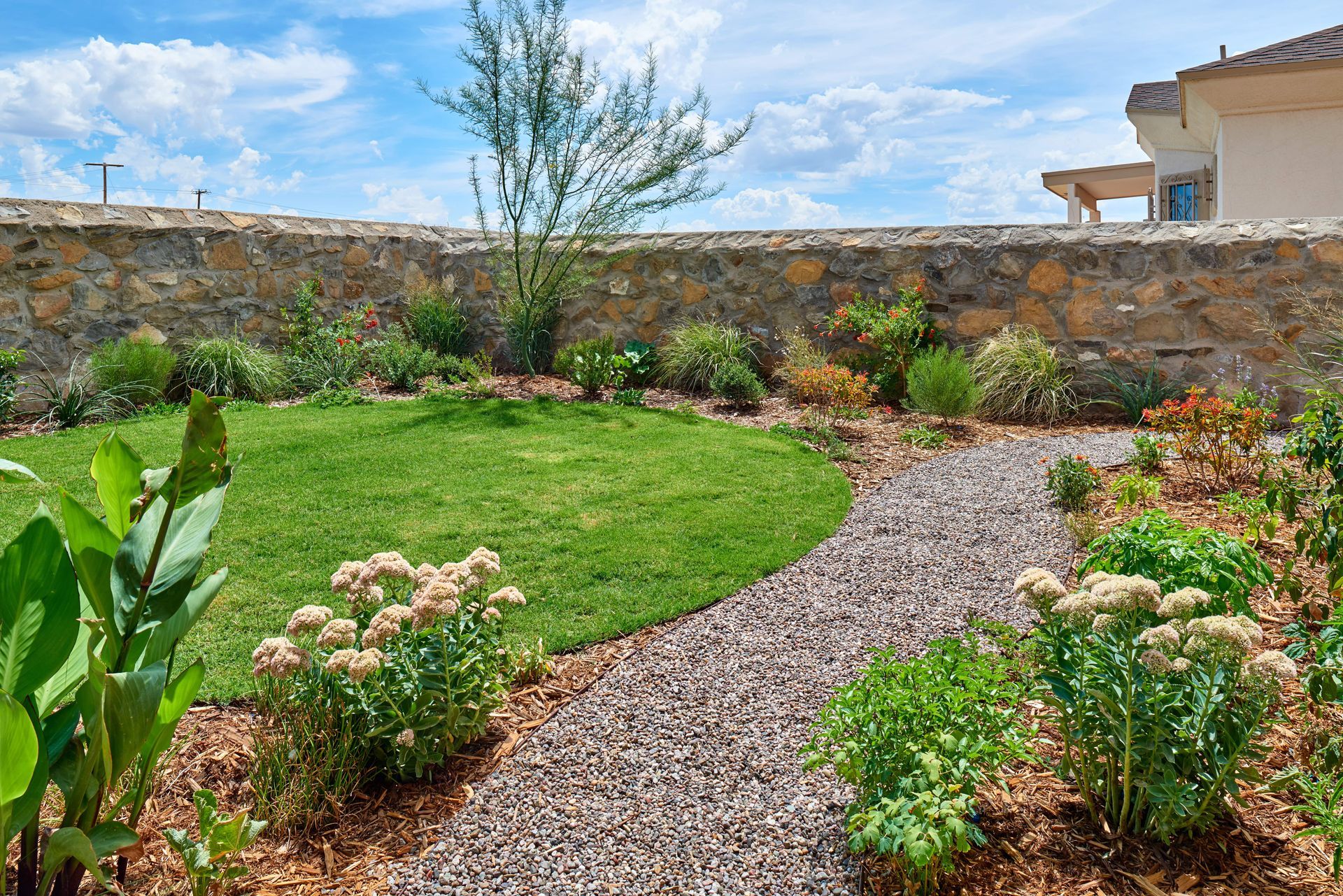 Garden with gravel path, green lawn, stone wall, and plants under a blue sky.