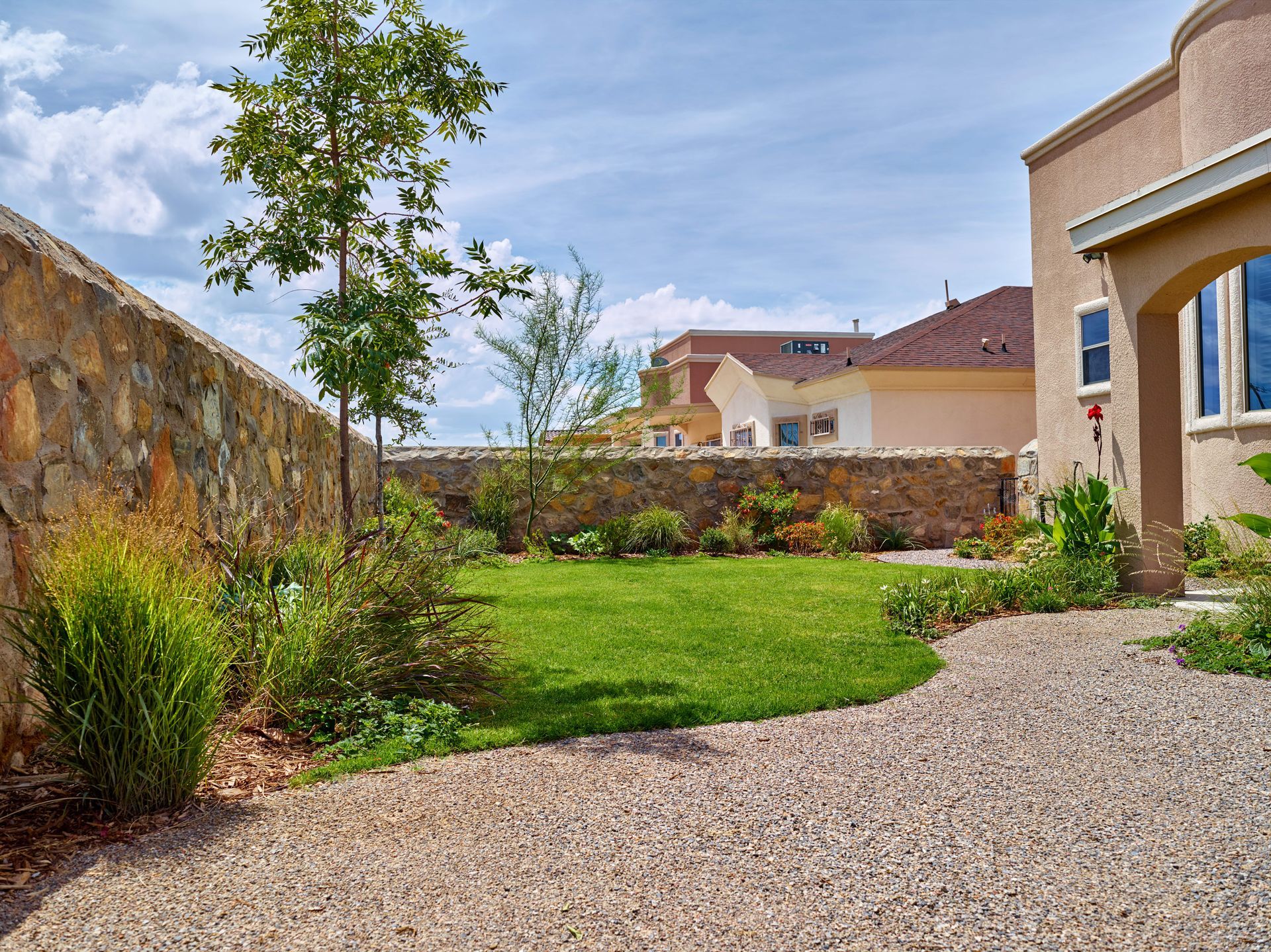 Stone-walled backyard with green lawn, gravel path, and plants, under a cloudy blue sky.