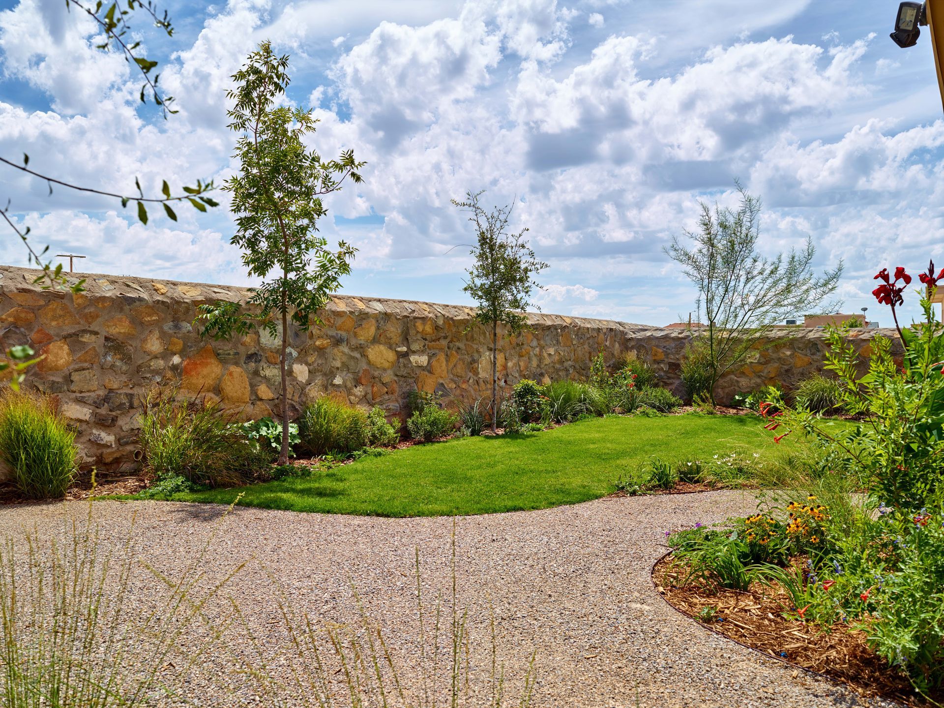 A gravel path leads through a garden with a small lawn, plants, and a stone wall under a cloudy sky.