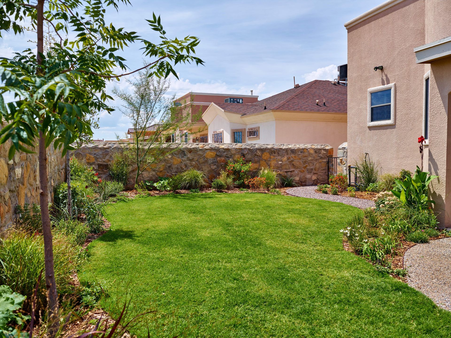 Green lawn in a backyard, bordered by stone wall and plants, with beige house exterior visible.