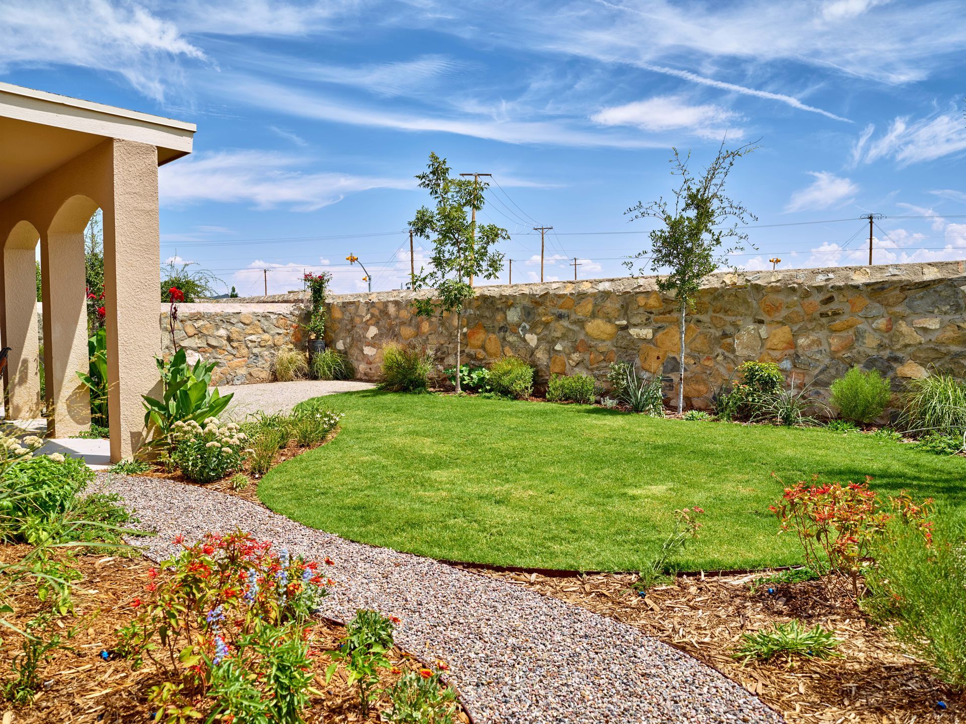 A lush green lawn surrounded by a stone path and flower beds, next to a building under a bright blue sky.
