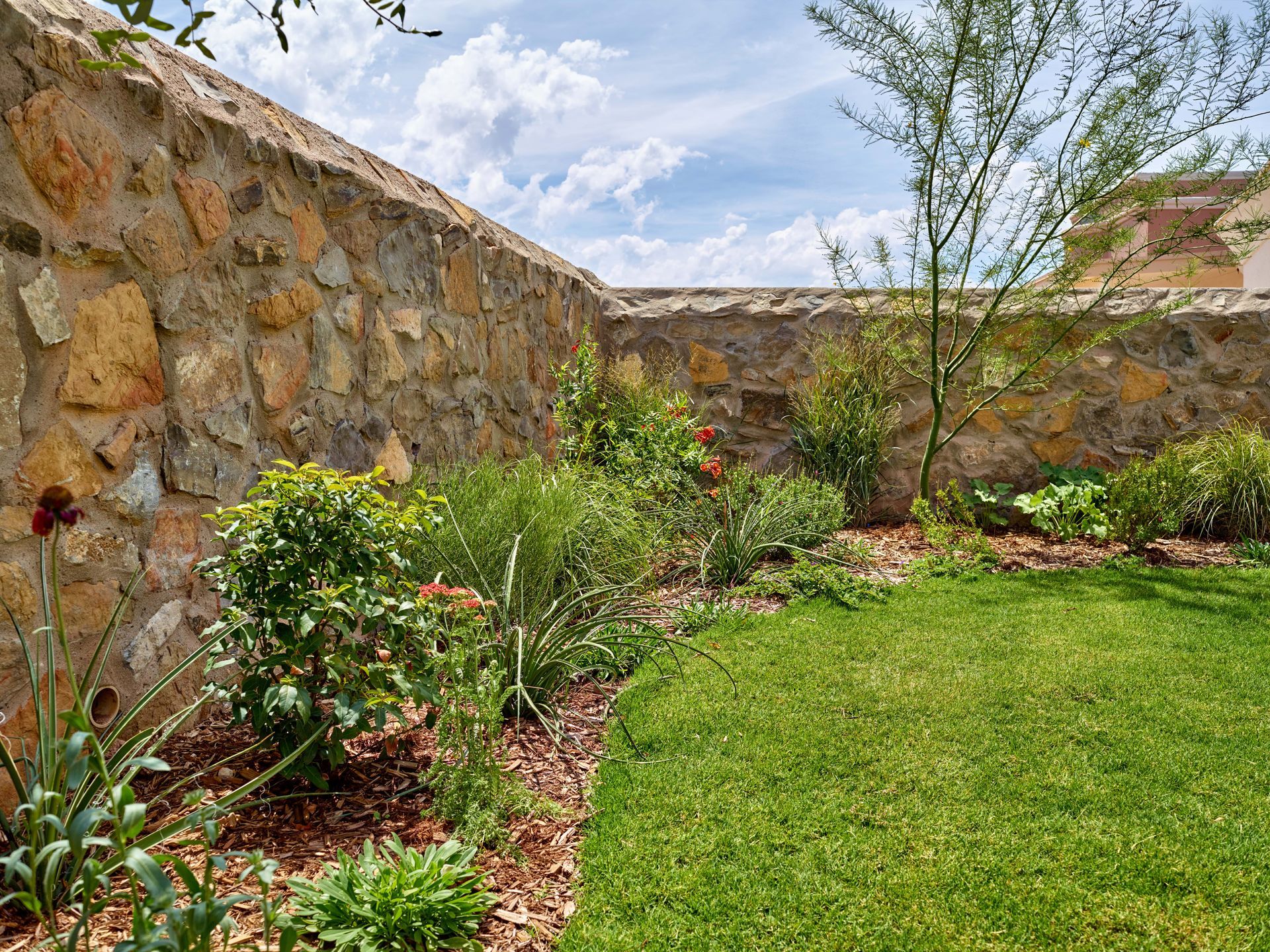 Stone wall surrounds a green lawn and garden beds with various plants under a cloudy sky.